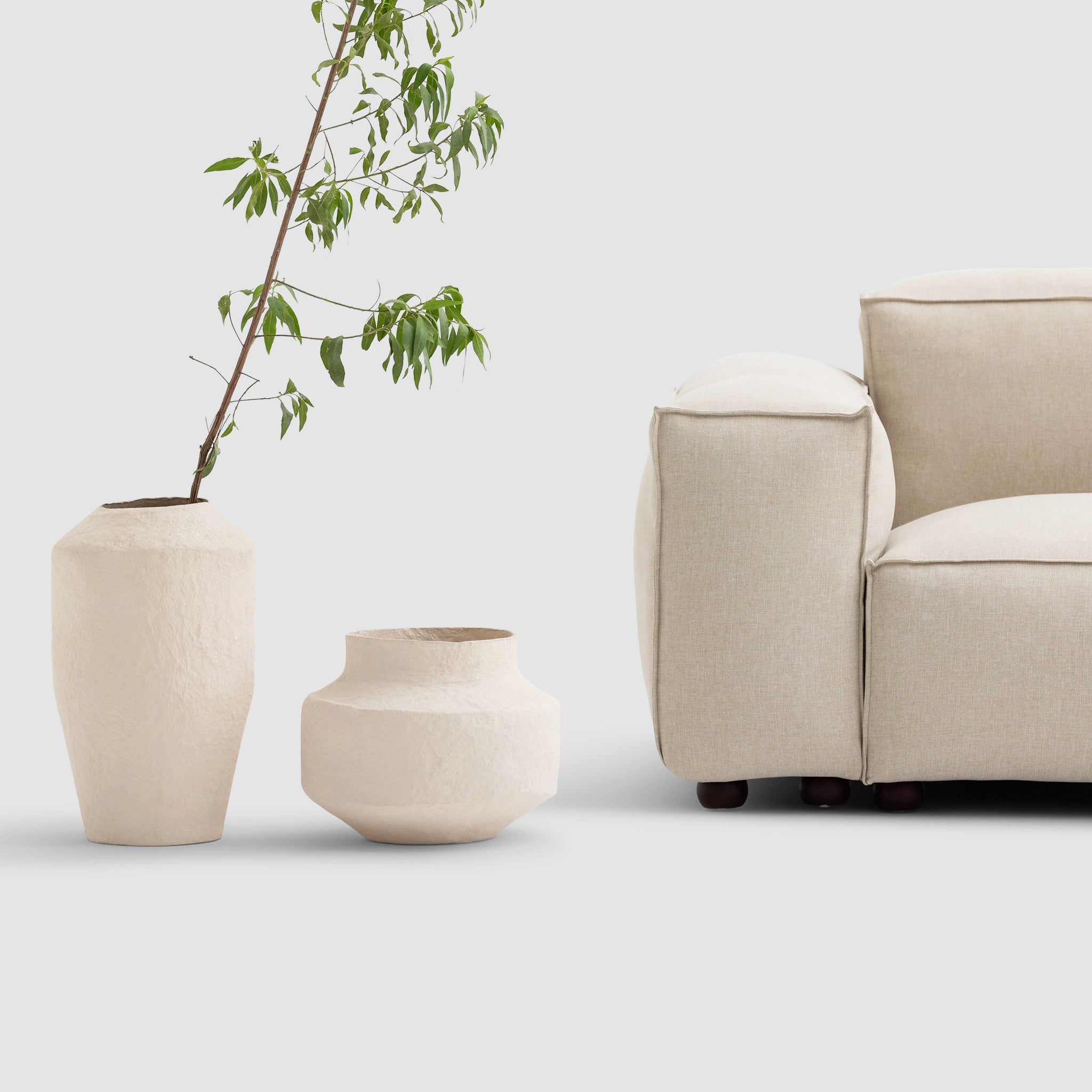 A detailed view of a beige sofa's armrest on the right, accompanied by two textured beige vases on the left. One vase is tall with a leafy branch, while the other is shorter and wider, both set against a light grey background.