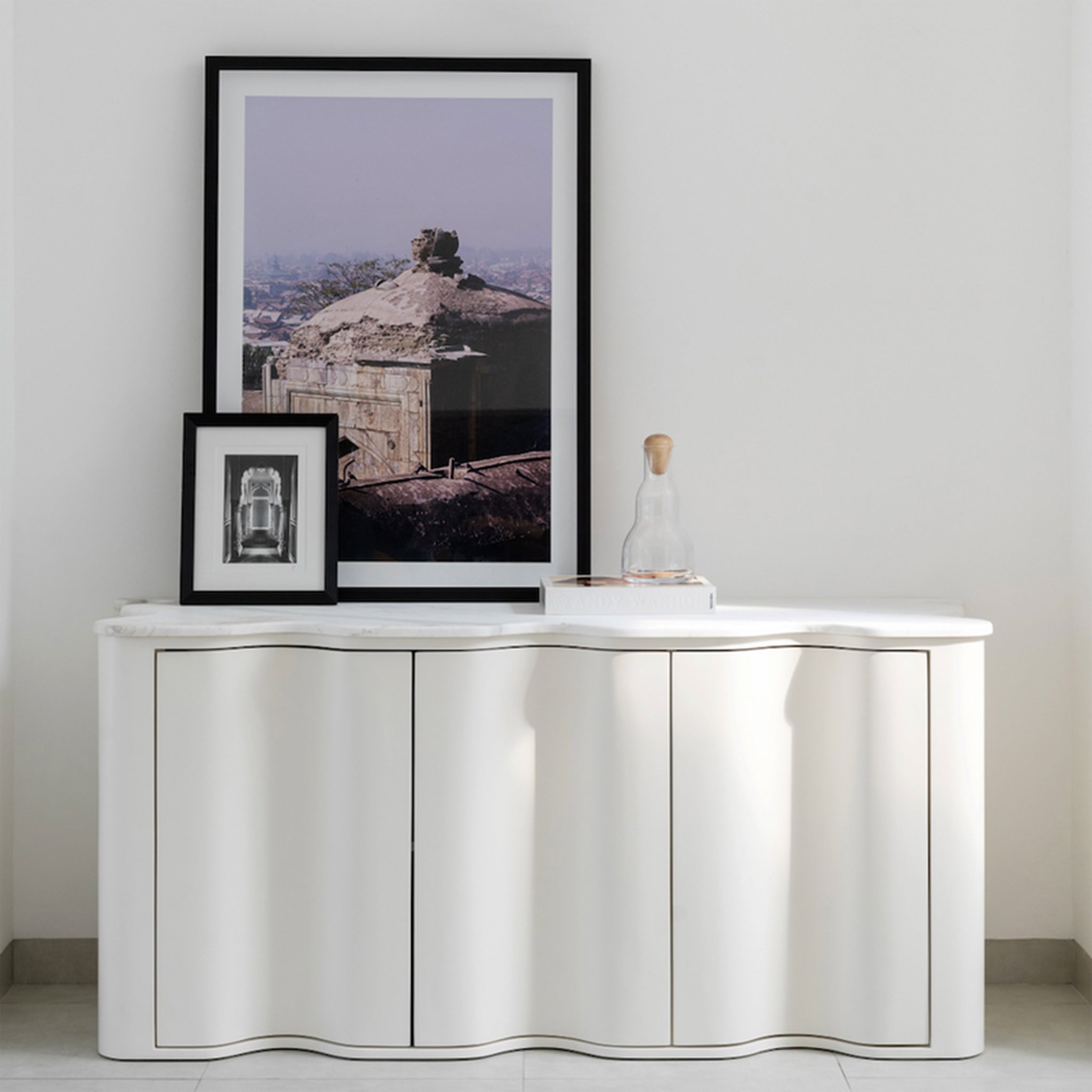 A chic sideboard with a wavy front design and a white marble top, adorned with framed photographs, a glass carafe, and a book. The sideboard is set against a light gray wall, creating a minimalist and elegant look in the living space.