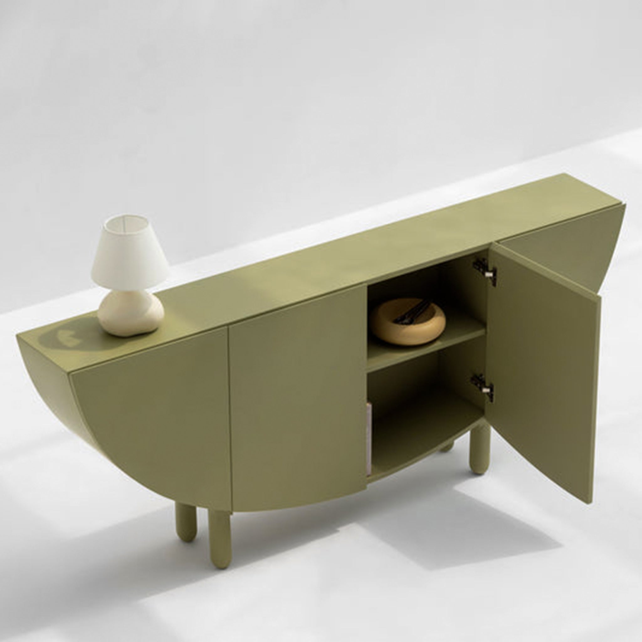 Top view of an olive green sideboard with a half-moon design, featuring an open compartment and a small white table lamp on top. One of the cabinet doors is open, revealing a bowl and other items inside, set against a light gray background.