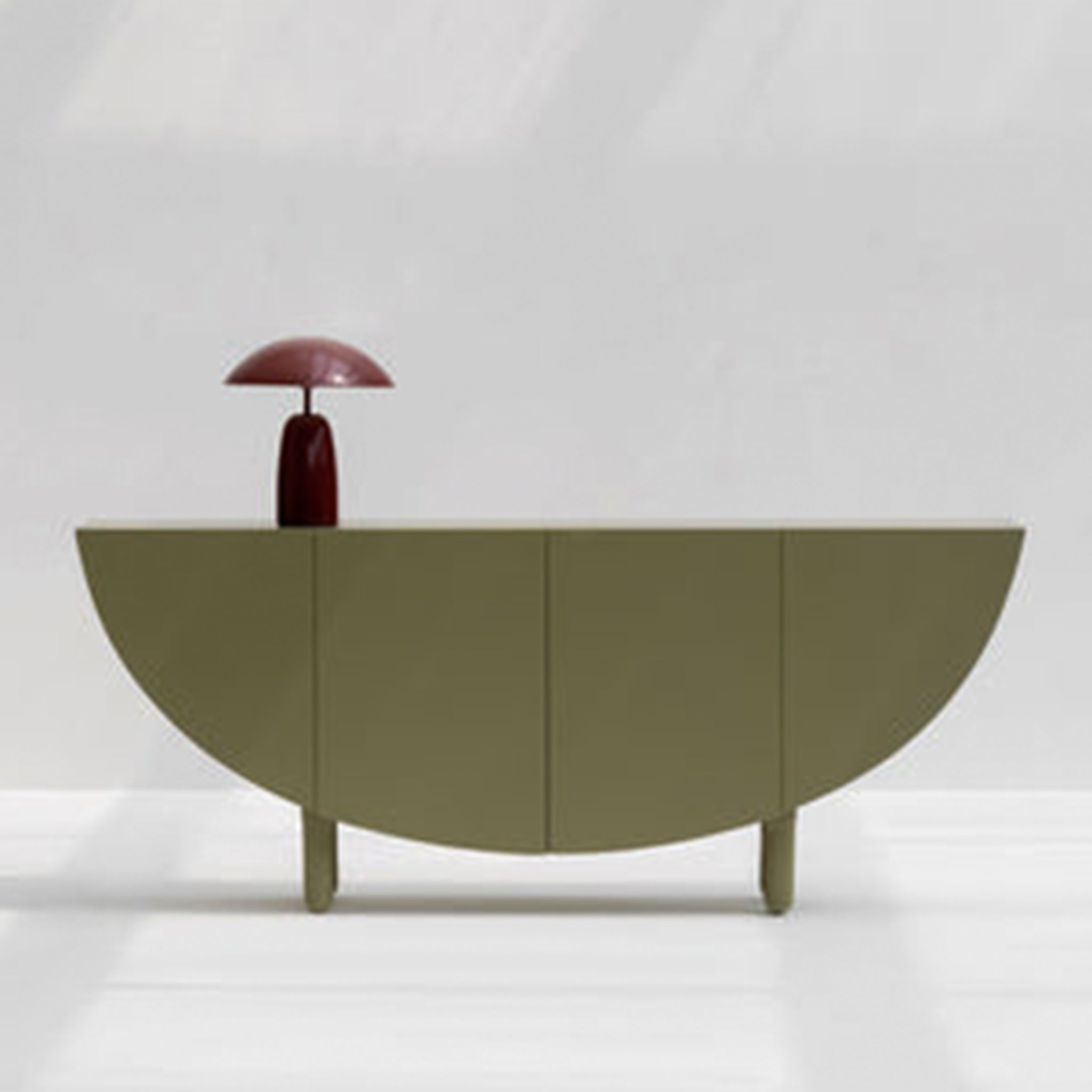A minimalist olive green sideboard with a half-moon design, featuring a burgundy table lamp on top, set against a light gray background.