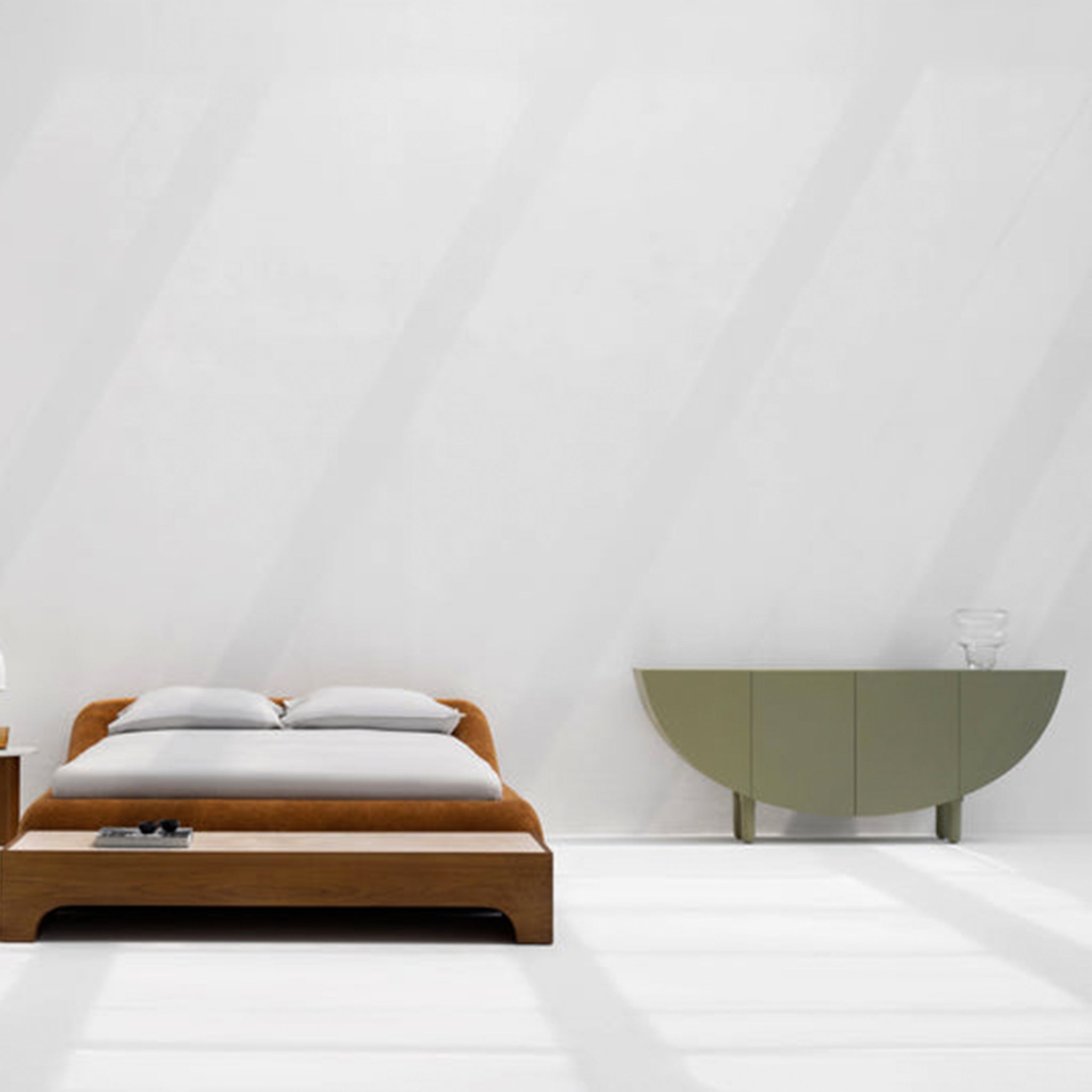 A minimalist bedroom featuring a wooden bed with white bedding, a small bedside table with a lamp, and an olive green half-moon sideboard against a light gray wall. The sideboard has a clear glass vase on top, creating a clean and modern look.