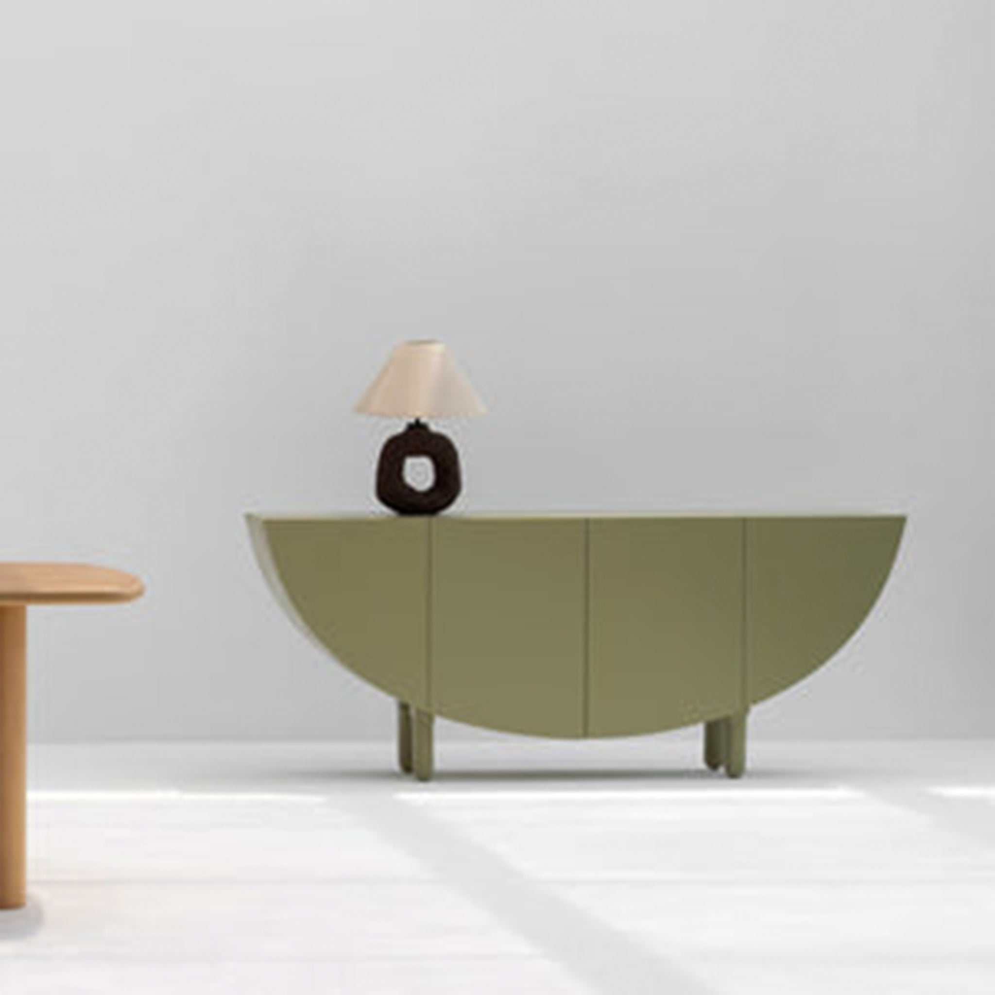 An olive green sideboard with a half-moon design, featuring a unique table lamp with a black base and beige shade, set against a light gray background. A portion of a wooden table is visible on the left side.