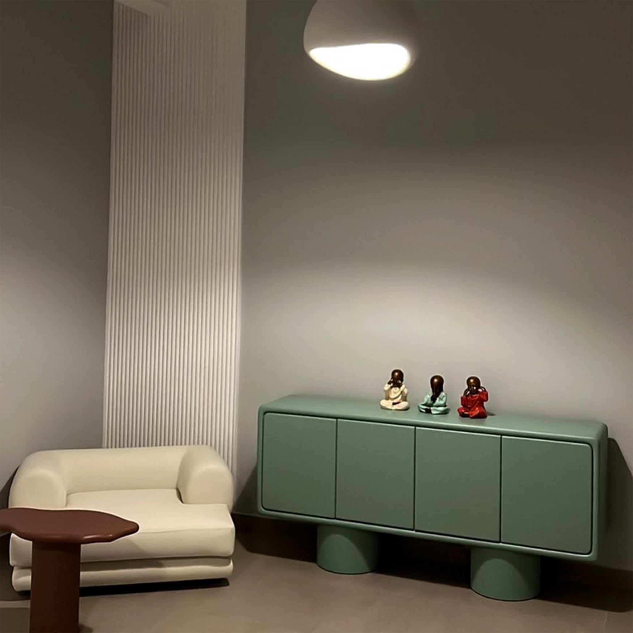 A modern interior featuring a green sideboard with four doors and cylindrical legs, adorned with decorative figurines on top, next to a white armchair and a unique brown side table, illuminated by a pendant light.