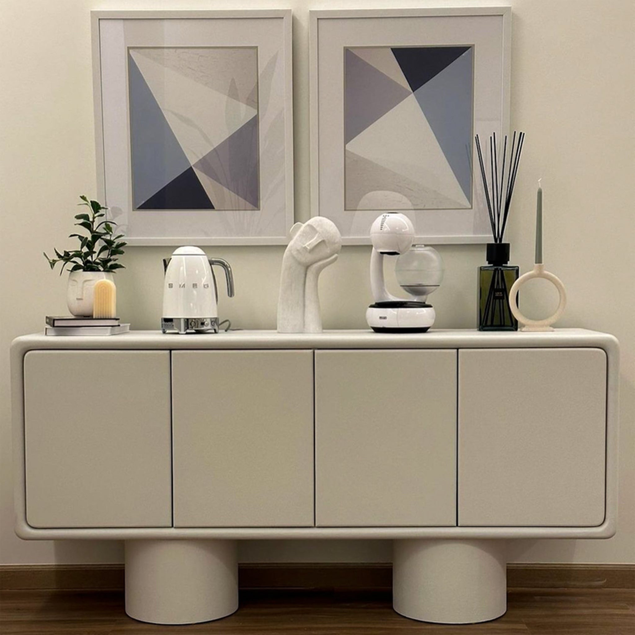 A beige sideboard with a minimalist design, featuring four doors and cylindrical legs, decorated with modern art pieces, a small plant, a kettle, and other contemporary items, placed against a wall with geometric framed artworks.