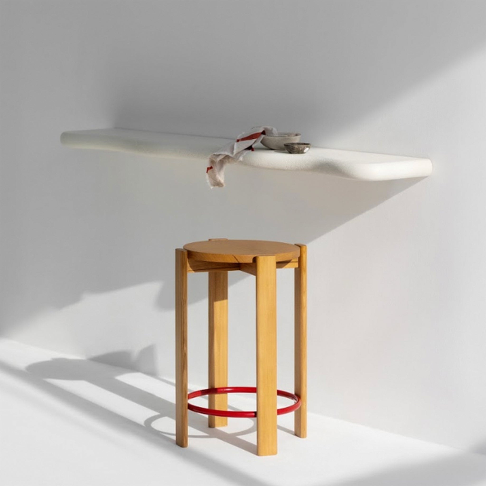 Wooden bar stool with a red circular footrest placed in a sunlit, minimalist kitchen with a floating white countertop