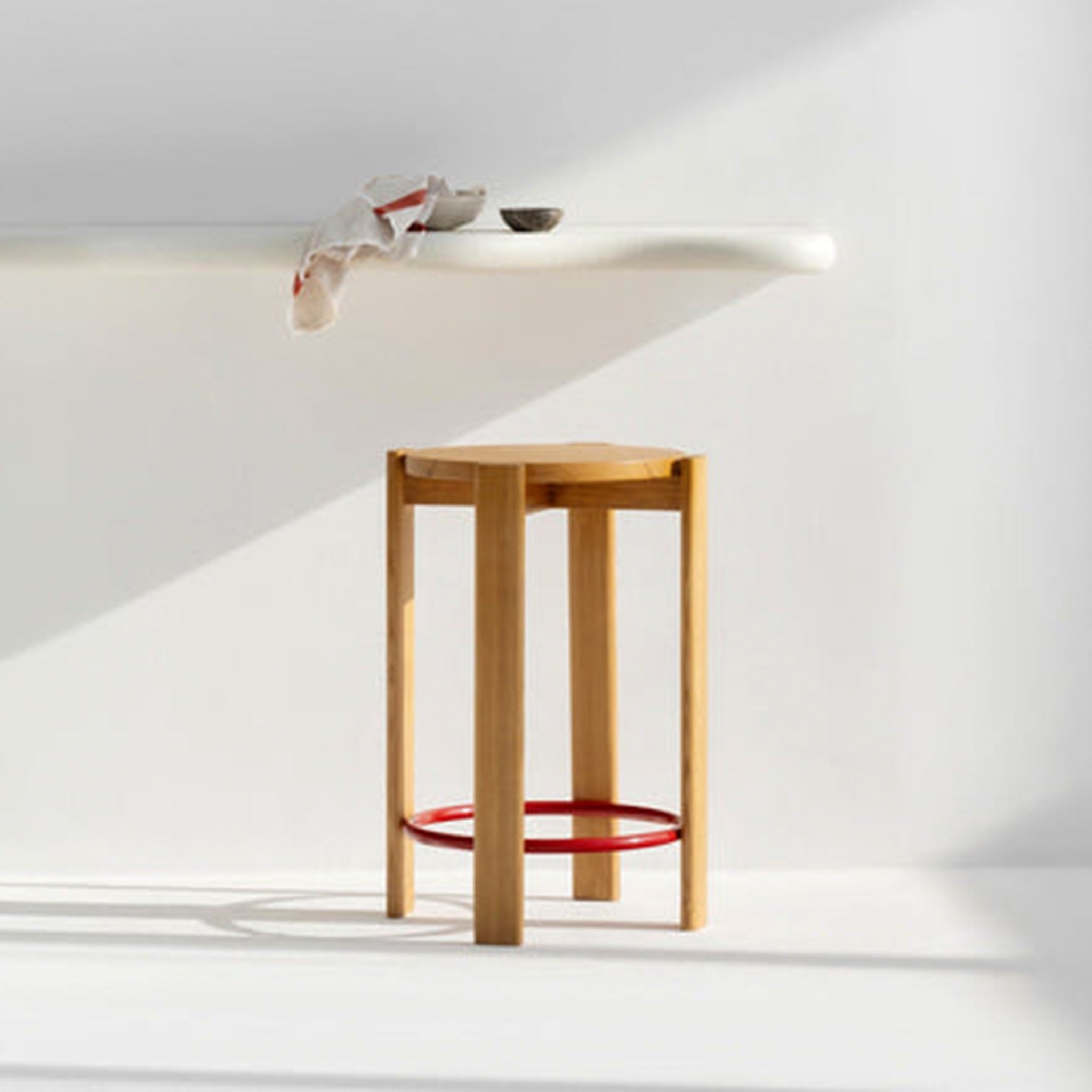 Wooden tall stool with a red circular footrest placed in a bright, minimalist kitchen with a floating white countertop