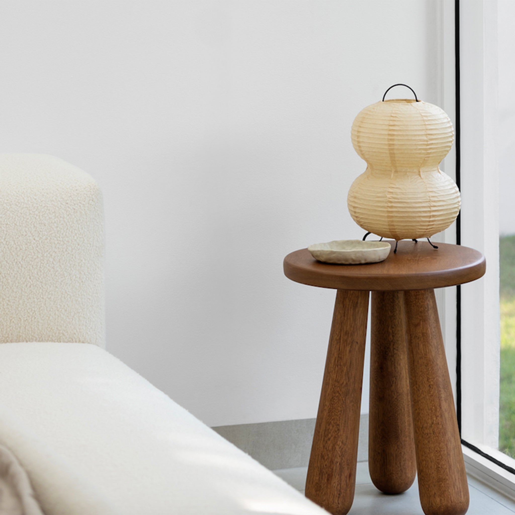 Wooden side table with chunky legs, decorated with a paper lantern and a small dish, next to a white sofa in a minimalist living room
