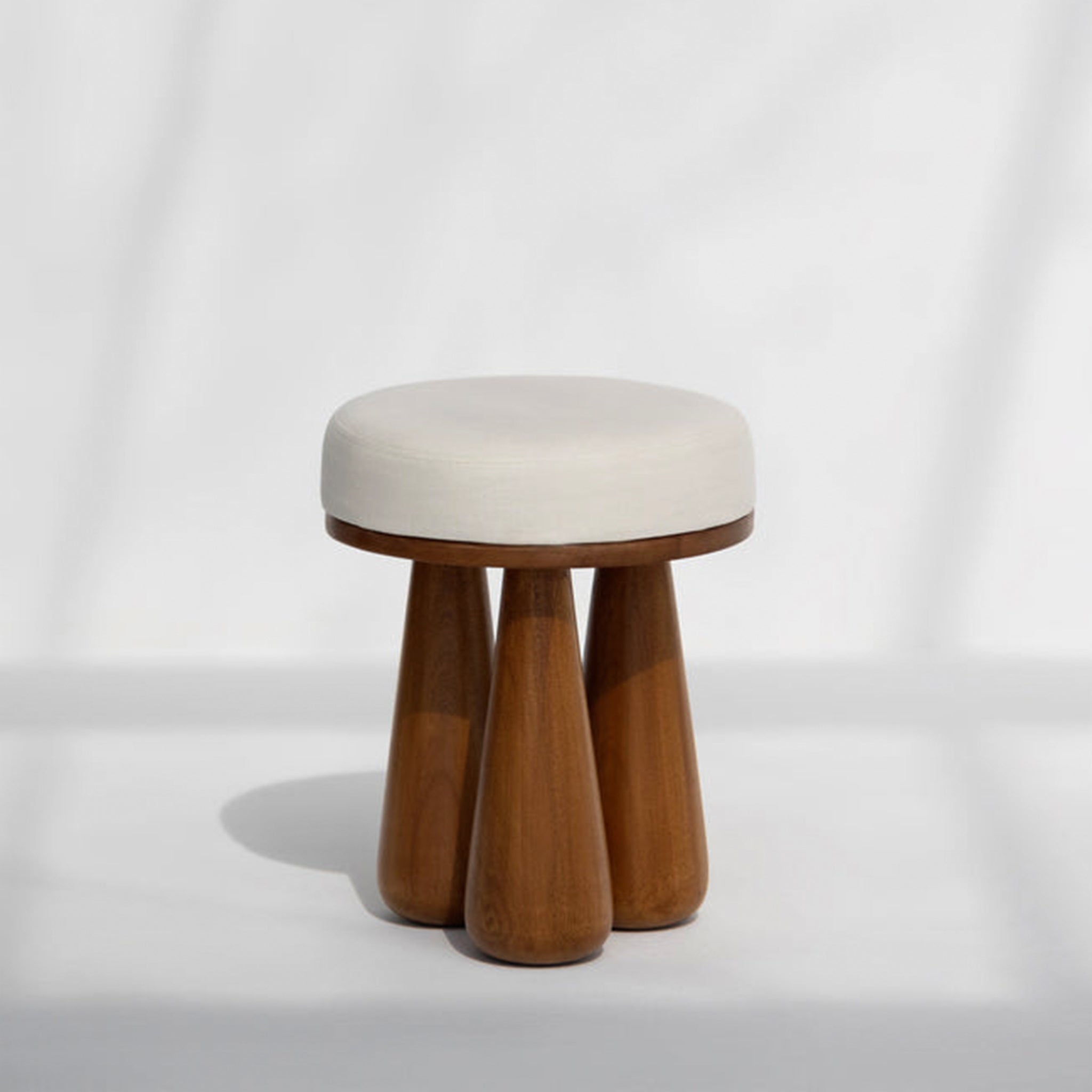 Wooden stool with chunky, tapered legs and a round, upholstered white seat in a sunlit room