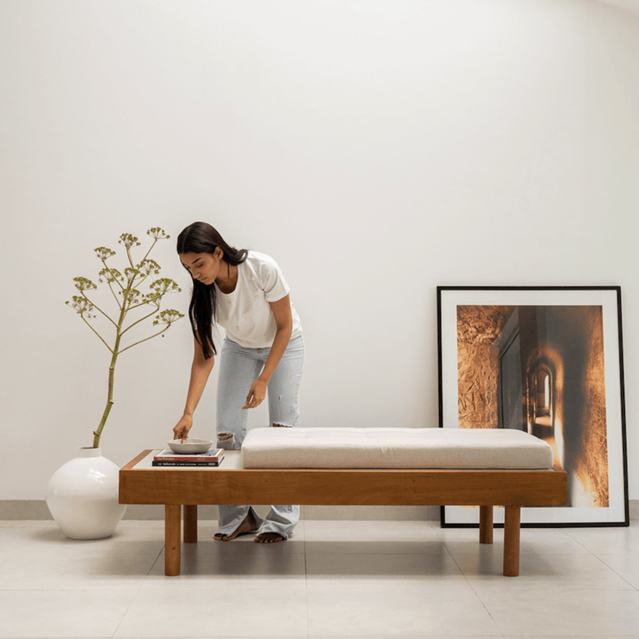 Woman arranging items on a modern wooden daybed with a customizable cushion and a side tray, in a minimalist room with a large vase and framed artwork