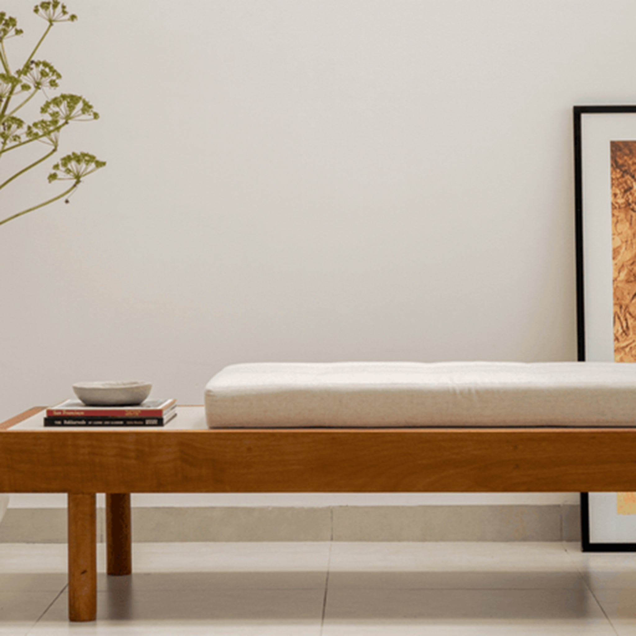 Modern wooden daybed with a customizable cushion and a side tray, placed in a minimalist room with a large vase and framed artwork