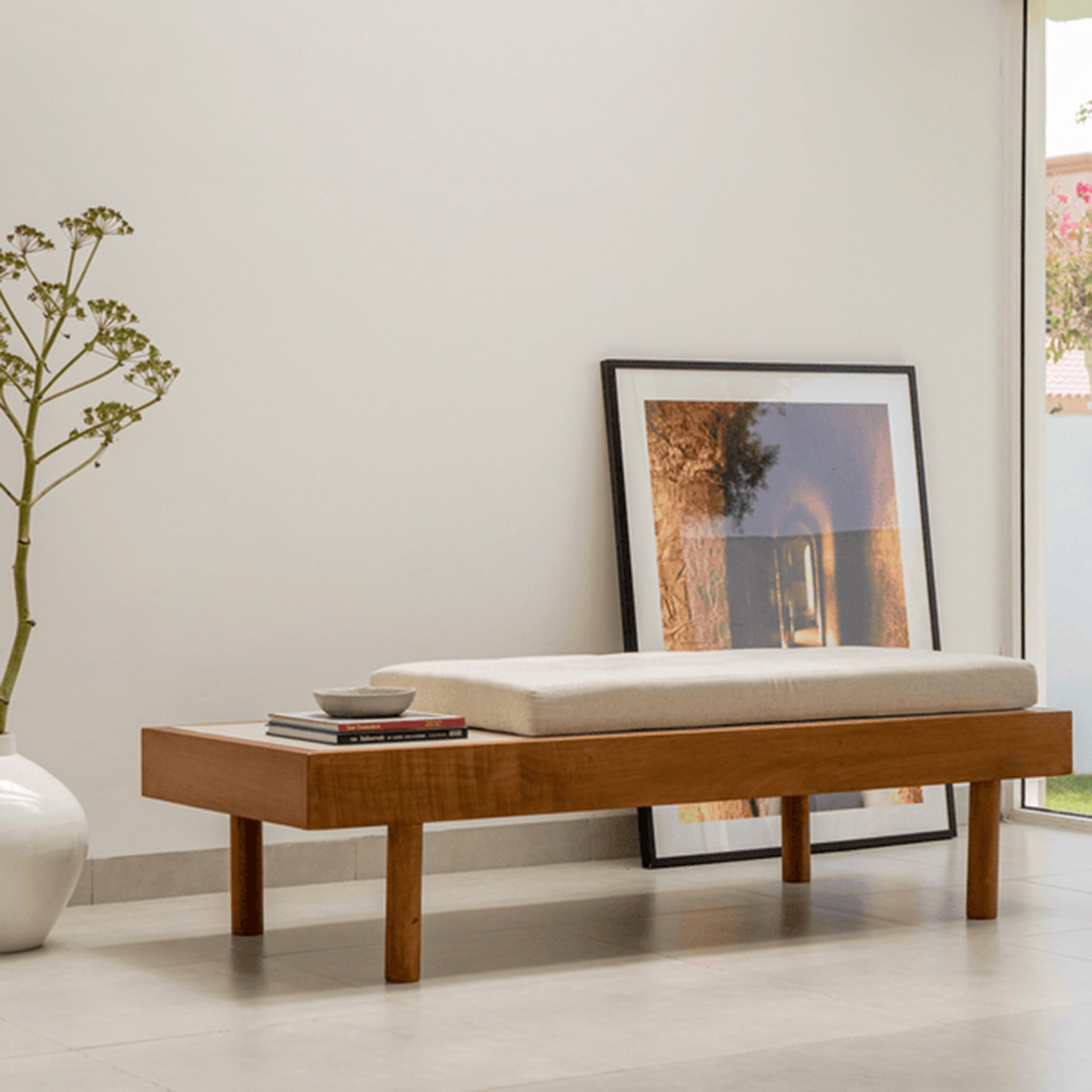 Modern wooden daybed with a customizable cushion and a side tray, placed in a bright minimalist room with a large vase and framed artwork leaning against the wall