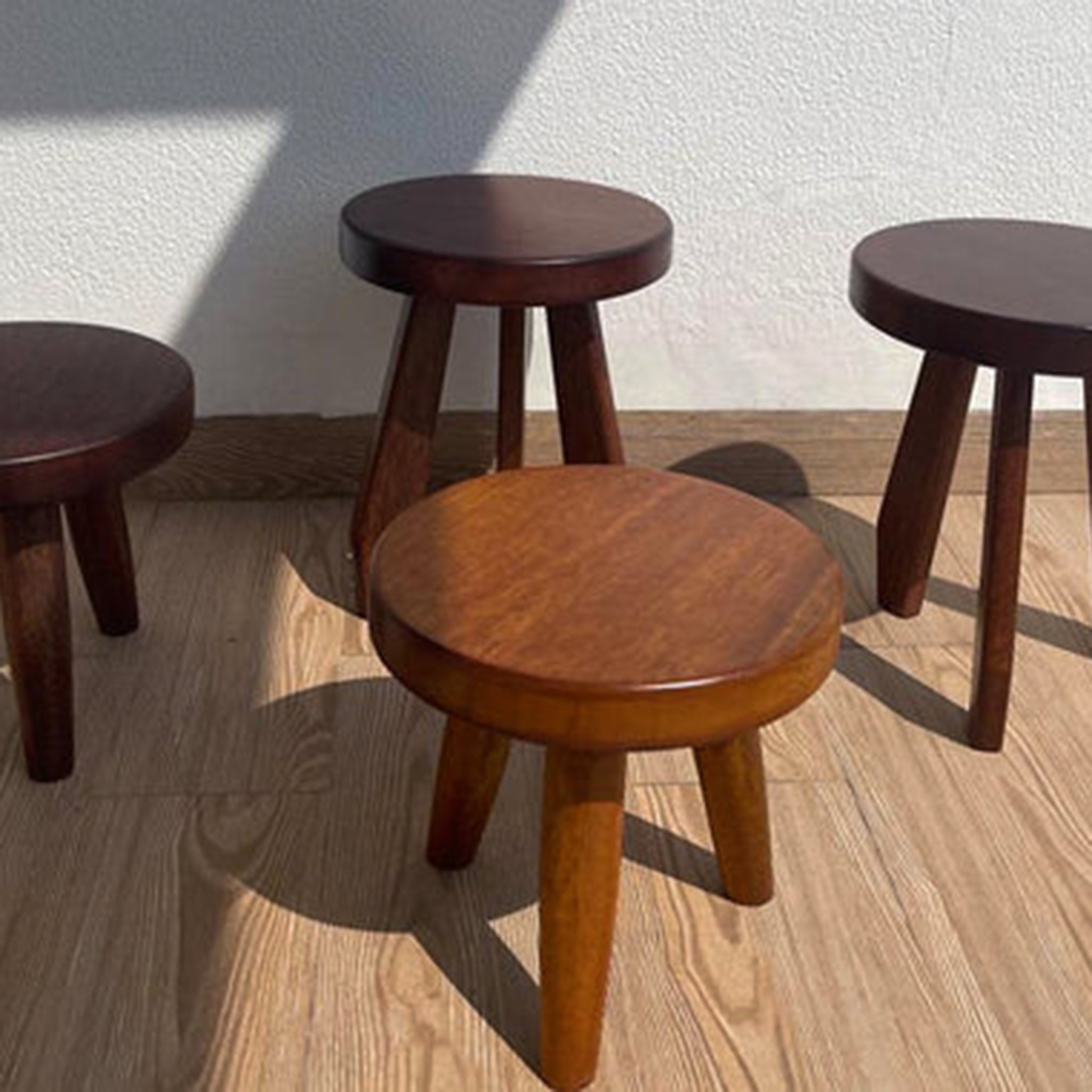 Four wooden three-legged mini stools in various shades of brown on a wooden floor
