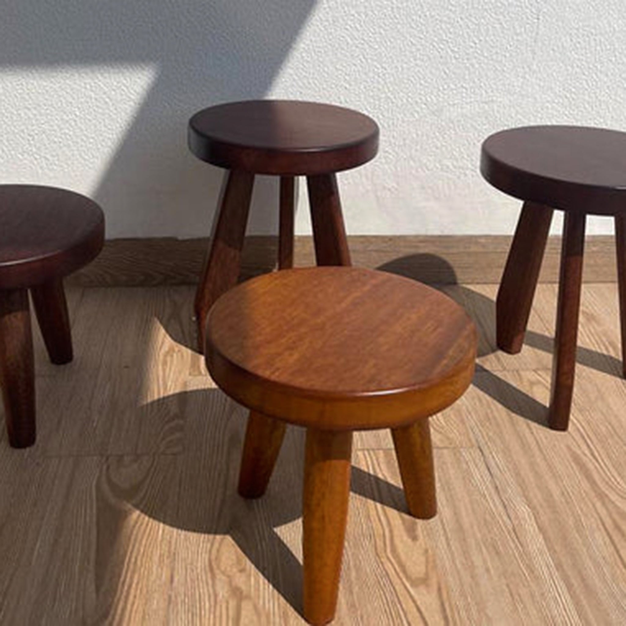 Four wooden three-legged stools in different shades of brown on a sunlit wooden floor