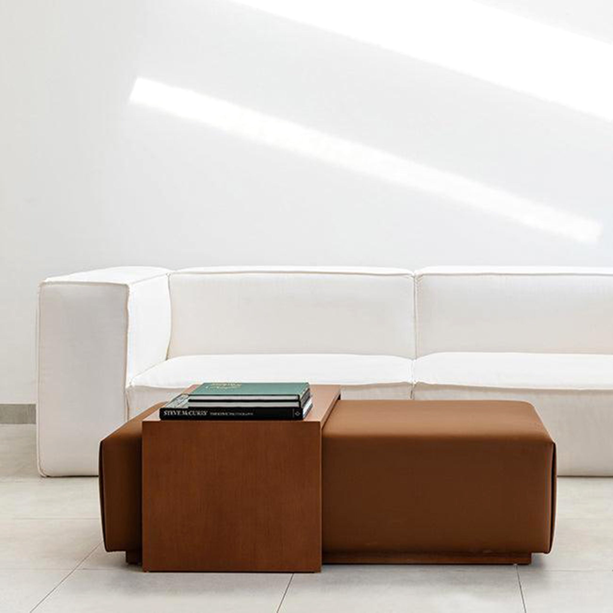 Modern living room with a white modular couch, accompanied by a brown leather ottoman and a wooden side table holding books, bathed in natural light.