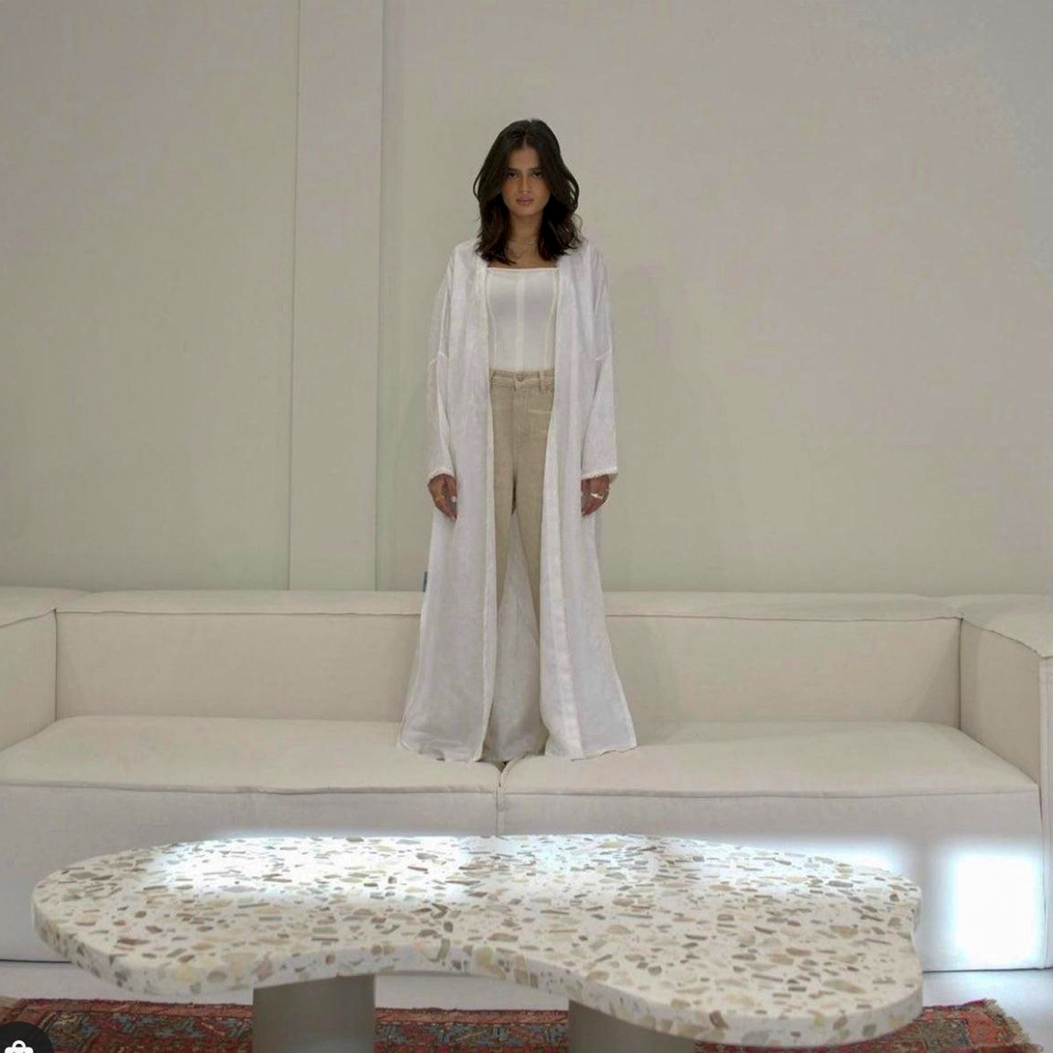 A woman in a white robe stands on a modern white modular couch, with a terrazzo coffee table in the foreground and a minimalist interior setting.