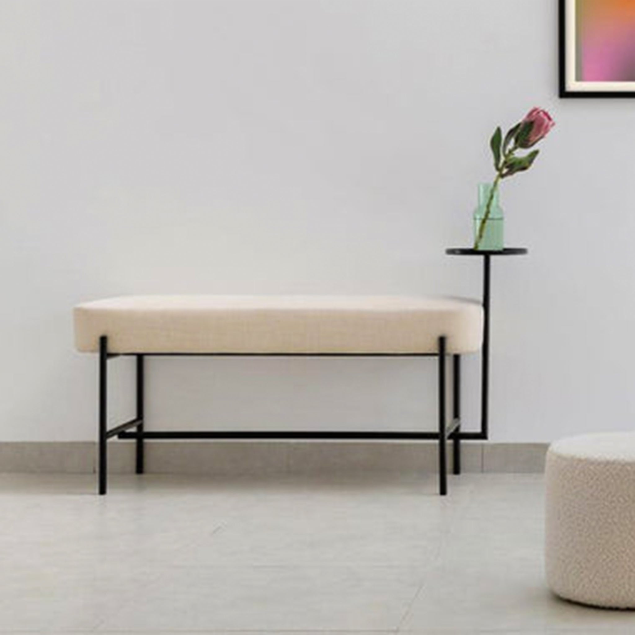 Modern bench with a beige upholstered seat and a matte black powder-coated steel frame, featuring an attached flat side table with a single rose in a glass vase, placed in a minimalist room