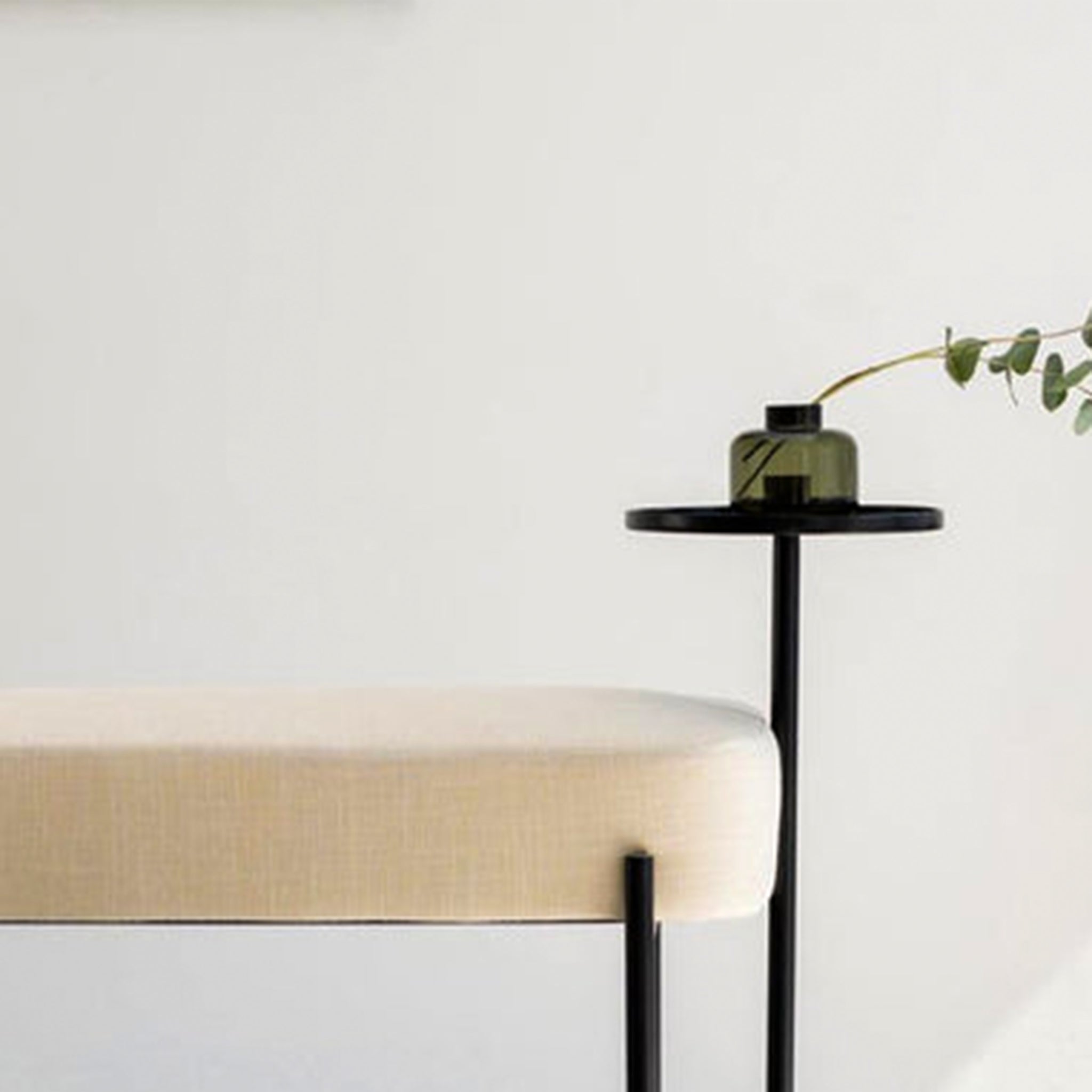 Close-up of a modern bench with a beige upholstered seat and a matte black powder-coated steel frame, featuring an attached flat side table with a green glass vase and eucalyptus branch