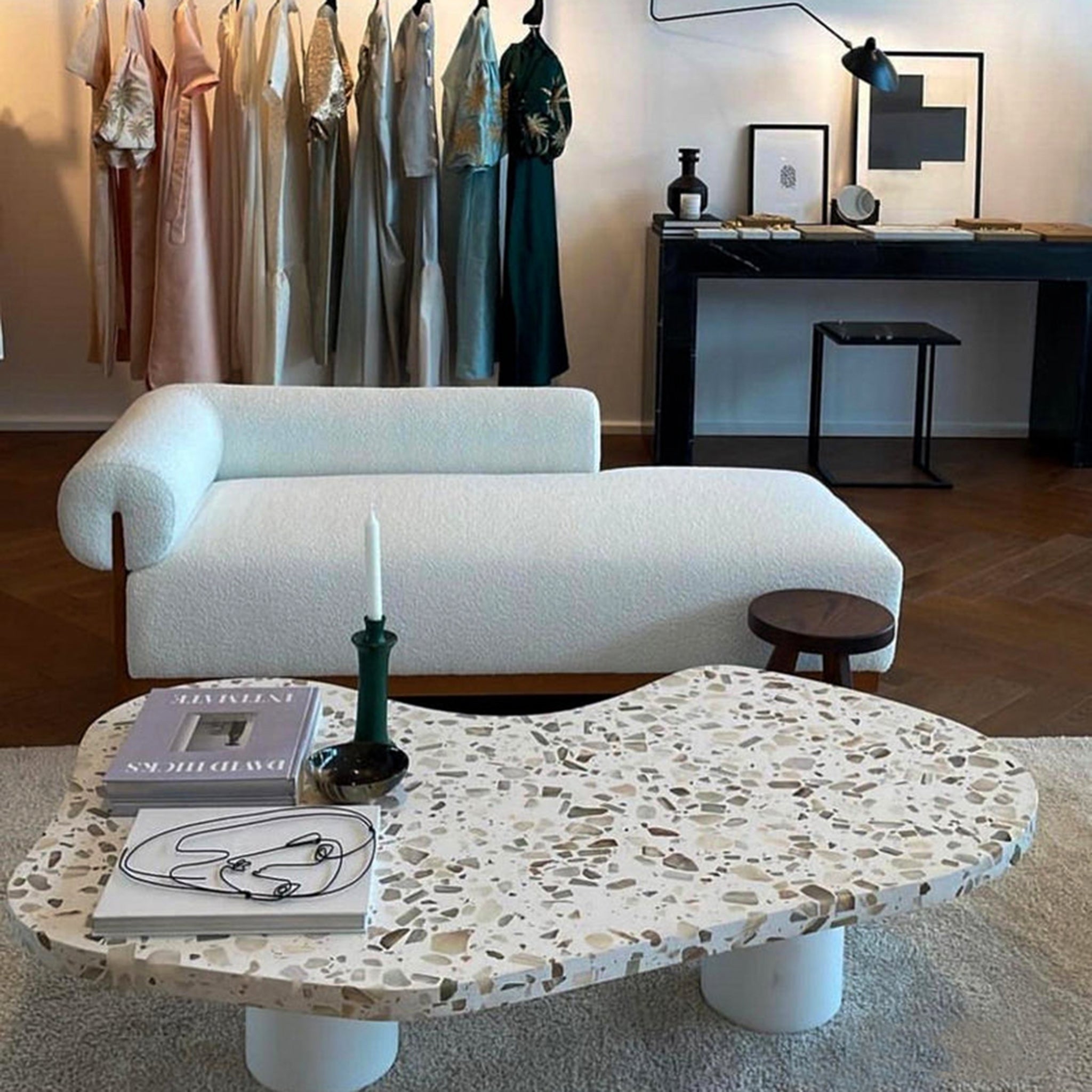 The Devon Chaise in a chic, modern interior with white upholstery and wooden frame, set near a unique terrazzo coffee table adorned with books and a candle, with a background of hanging clothes and a stylish black desk.