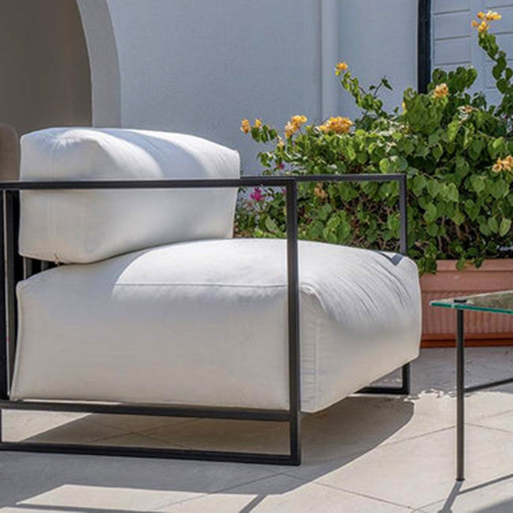 Outdoor setup featuring The Dexter Accent Chair with white cushions and a sleek black metal frame, placed on a patio with greenery and a glass side table.
