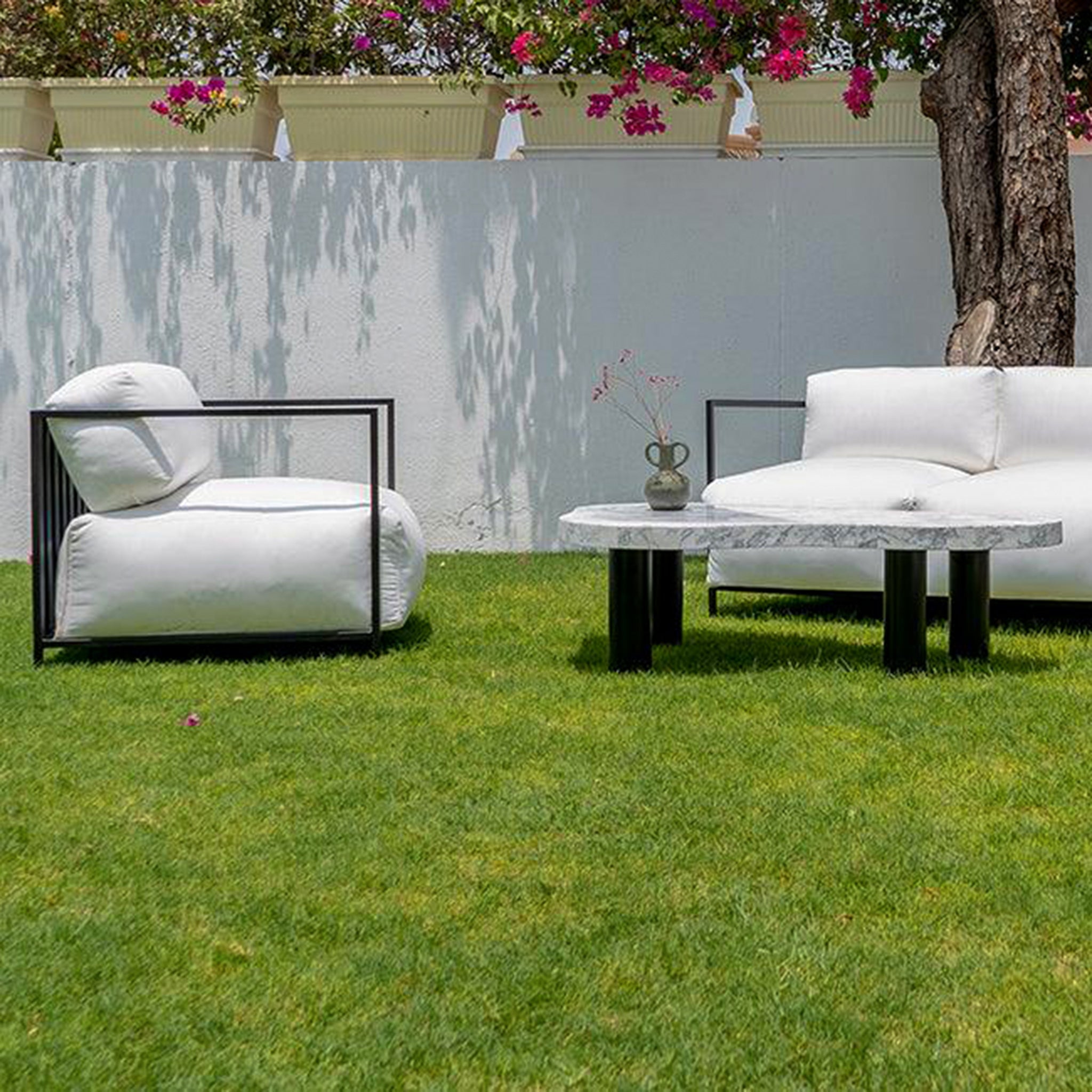 Outdoor garden setup featuring The Dexter Accent Chair with white cushions and black metal frame, paired with a matching sofa and a marble coffee table on a lush green lawn.