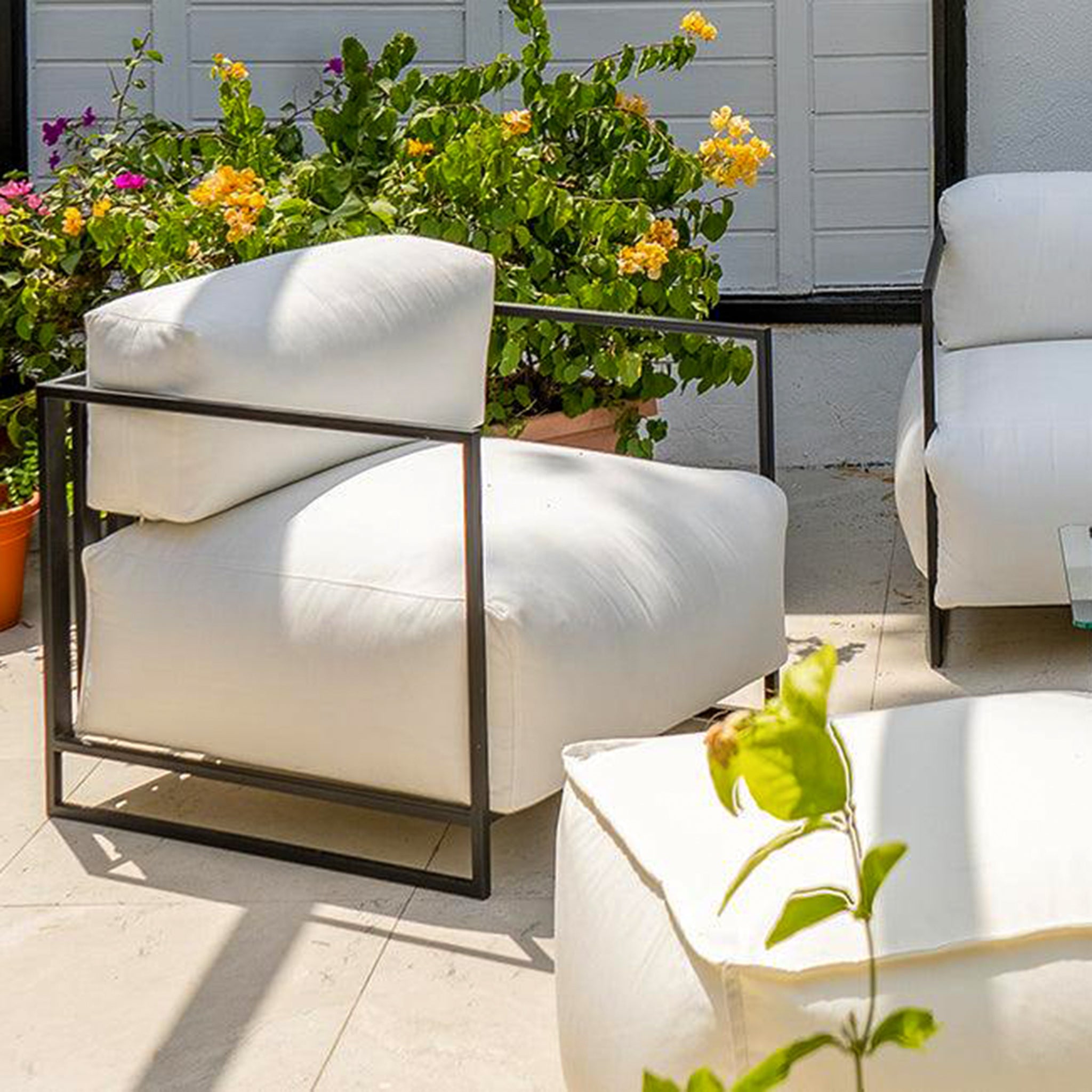 Sunny patio scene featuring The Dexter Accent Chair with white cushions and black metal frame, surrounded by vibrant potted plants and greenery.