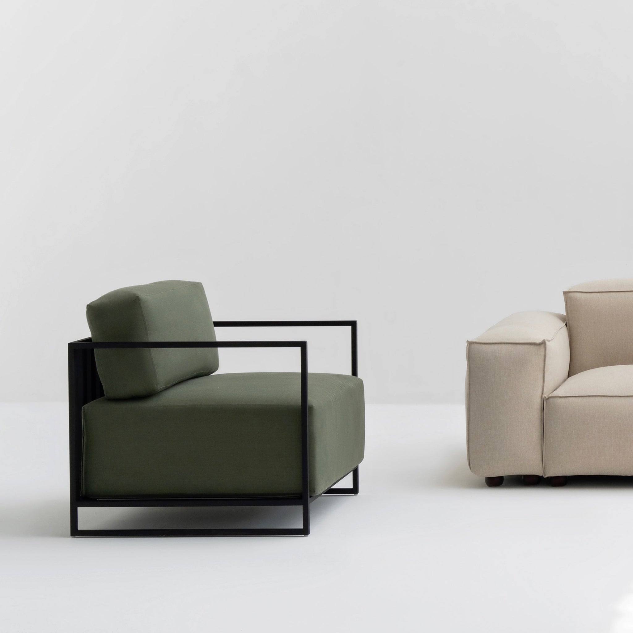 Modern interior featuring The Dexter Accent Chair with olive green cushions and a sleek black metal frame, positioned next to a beige sofa on a white background.