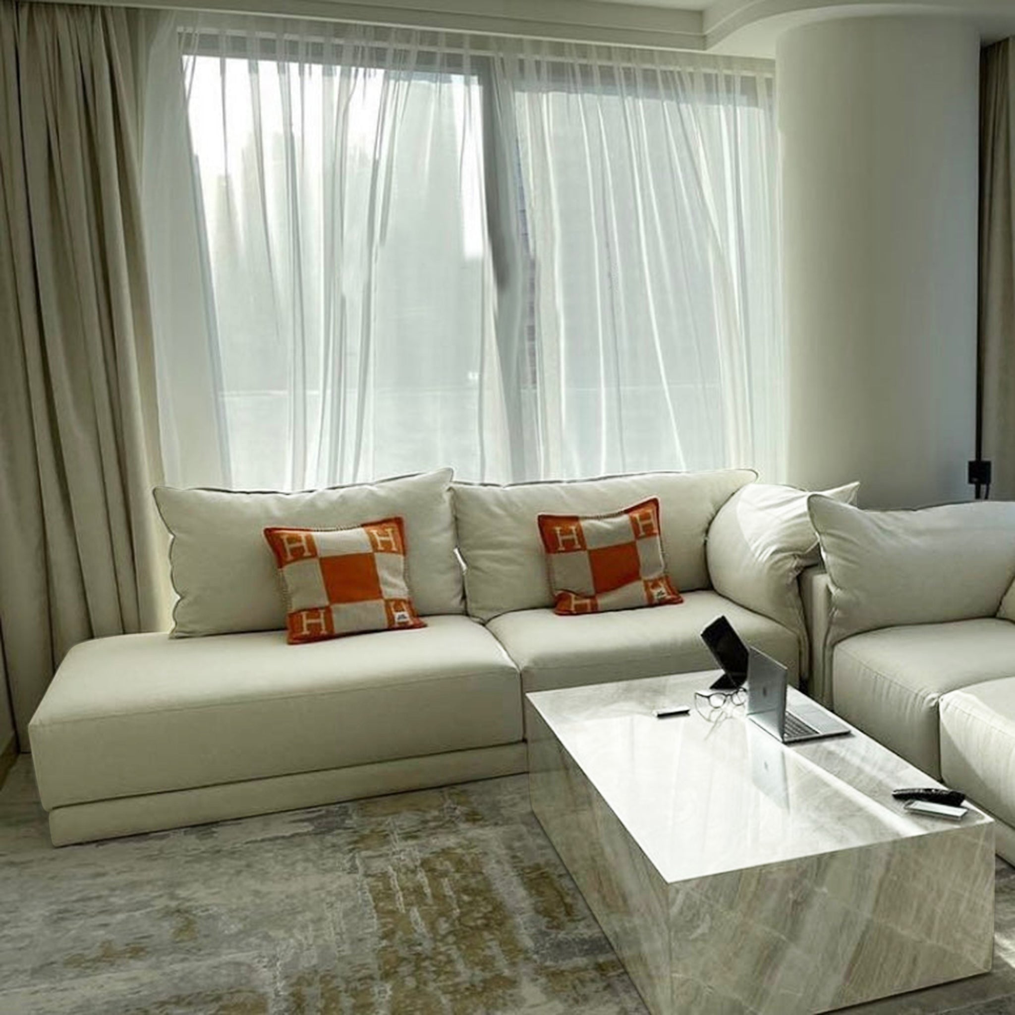 Elegant living room setup featuring a modern beige sectional sofa with plush cushions and orange accent pillows, complemented by a sleek marble coffee table and natural light streaming through sheer curtains.