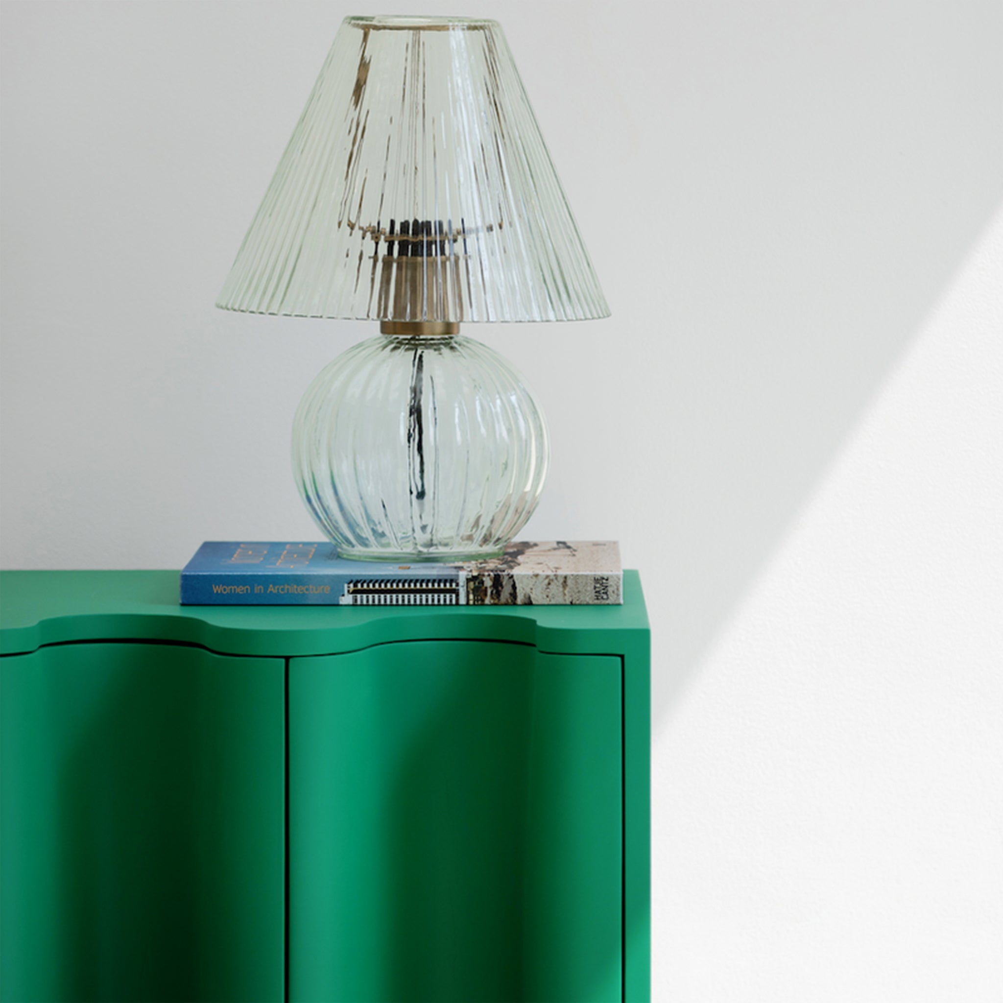 Close-up of a green bedside table with a wavy front design, featuring a clear glass table lamp and a stack of books on top, set against a white wall.