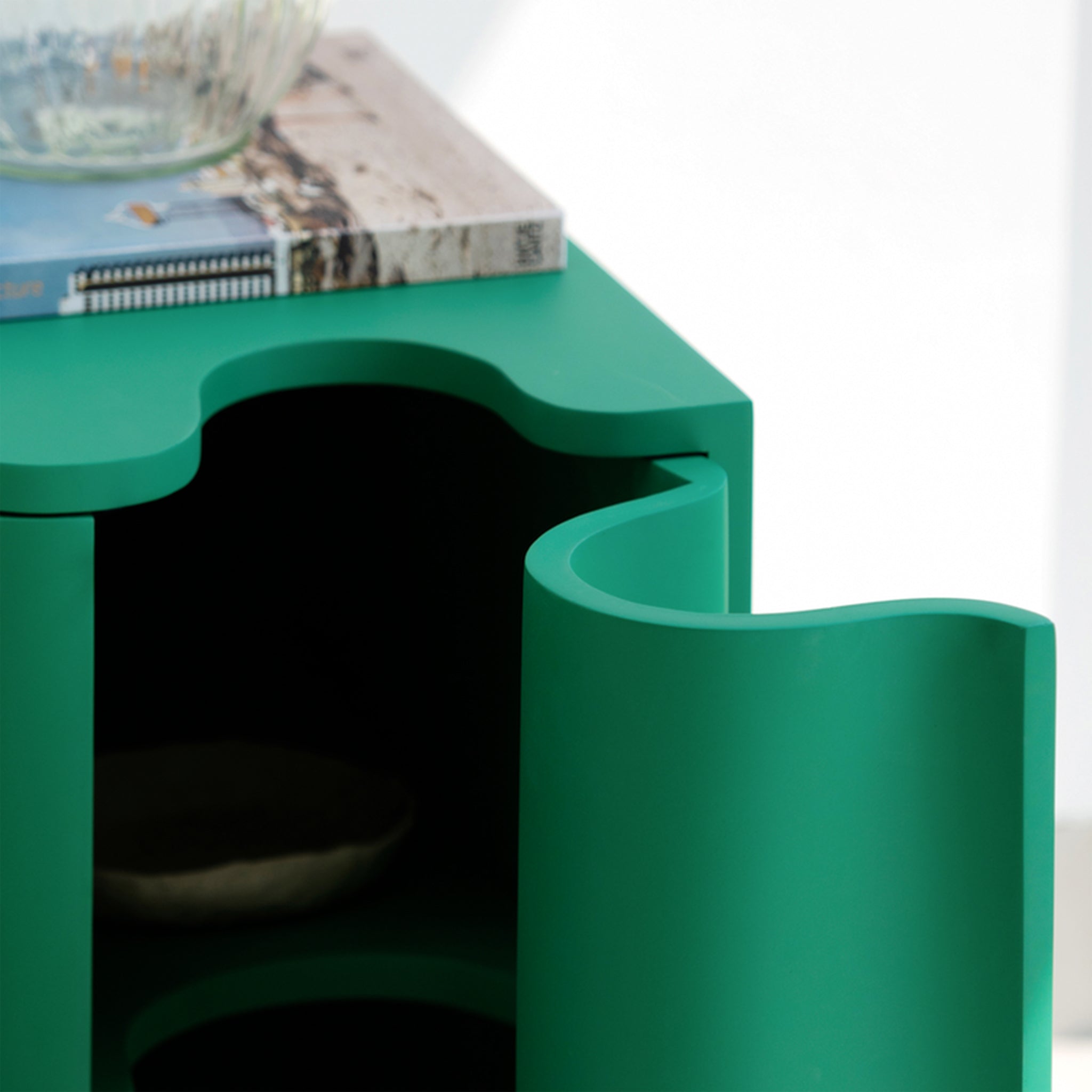 Close-up of a green bedside table with a wavy front design, showing an open compartment with a clear glass table lamp and books on top, set against a white background.