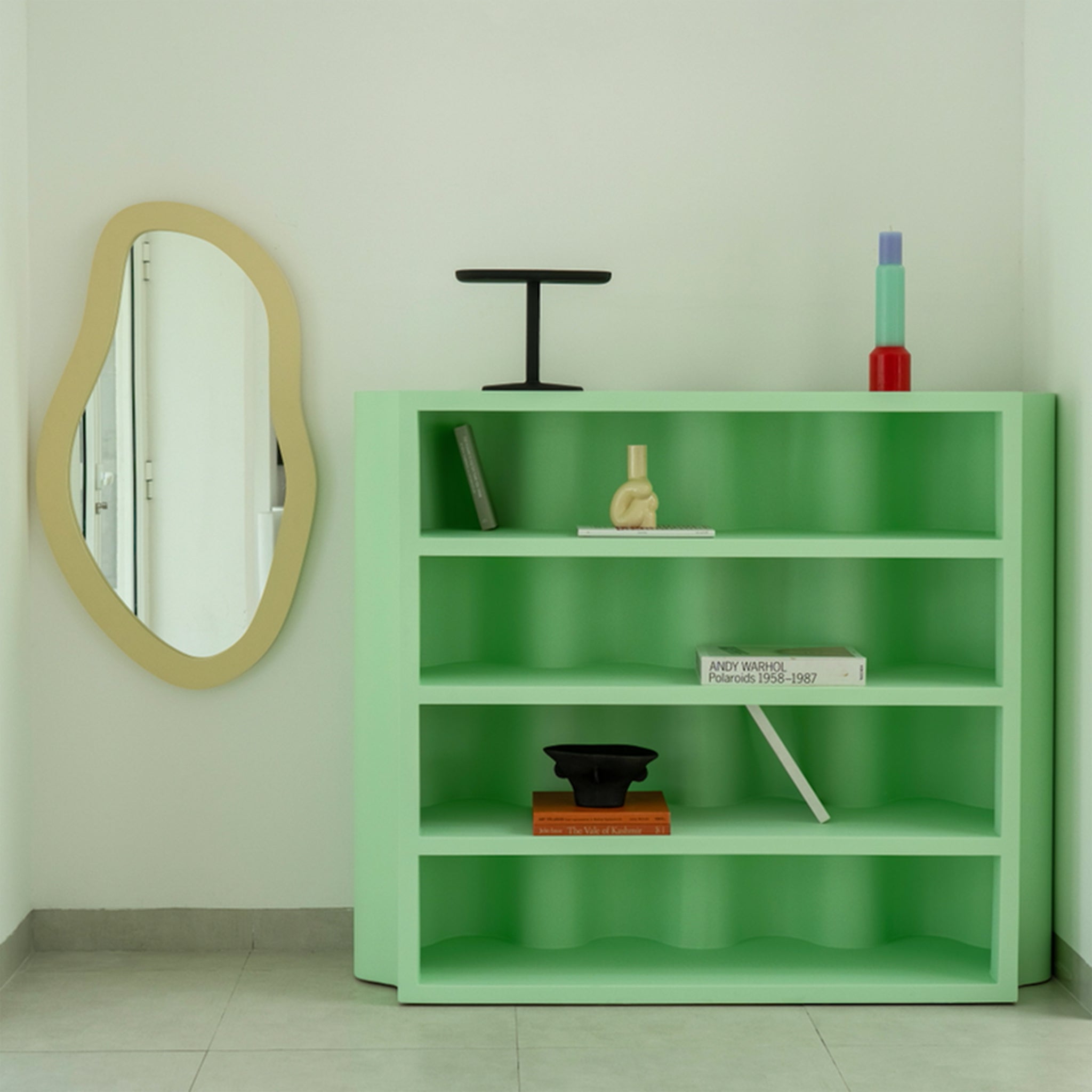 A contemporary green bookcase with a minimalist design, adorned with various decorative items and a book, placed next to a uniquely shaped mirror on the wall.