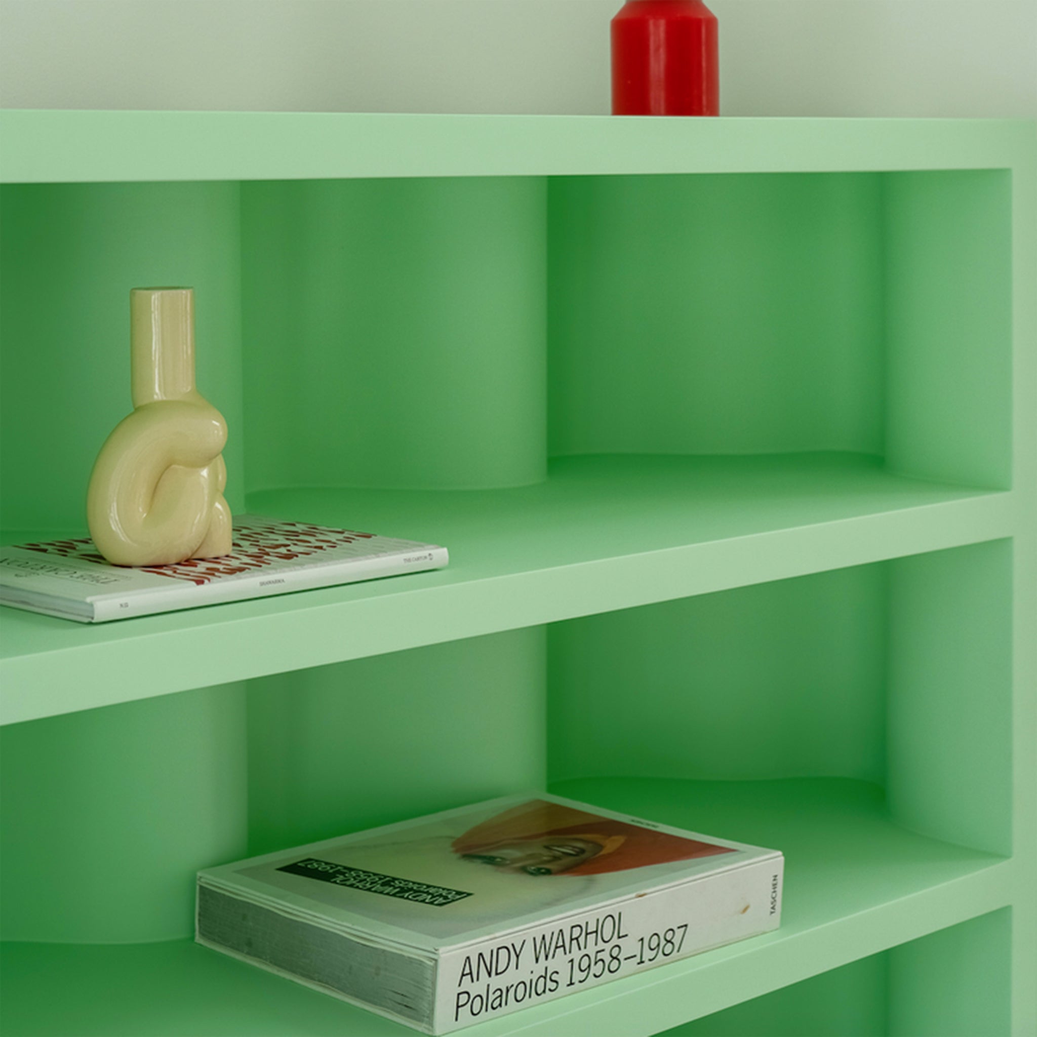 Close-up of a modern green bookcase displaying a unique ceramic sculpture, a red candle, and an Andy Warhol book.