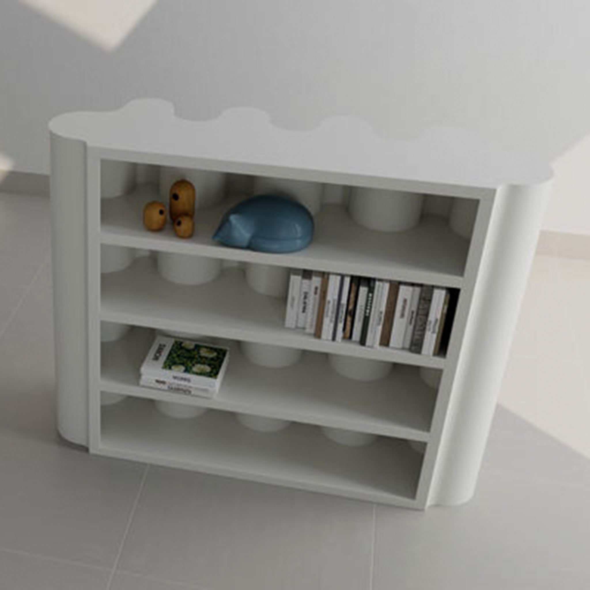 A modern white bookcase with a wavy top and rounded edges, featuring a collection of books and decorative items, including a blue ceramic cat and wooden sculptures.