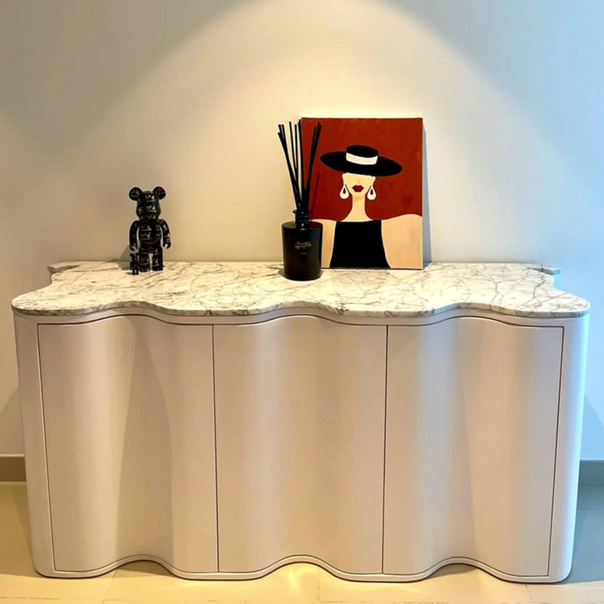 A stylish sideboard with a wavy front design and a white marble top, decorated with a black figurine, a reed diffuser, and a modern art painting of a woman in a hat. The sideboard is placed against a light gray wall in a contemporary living space.