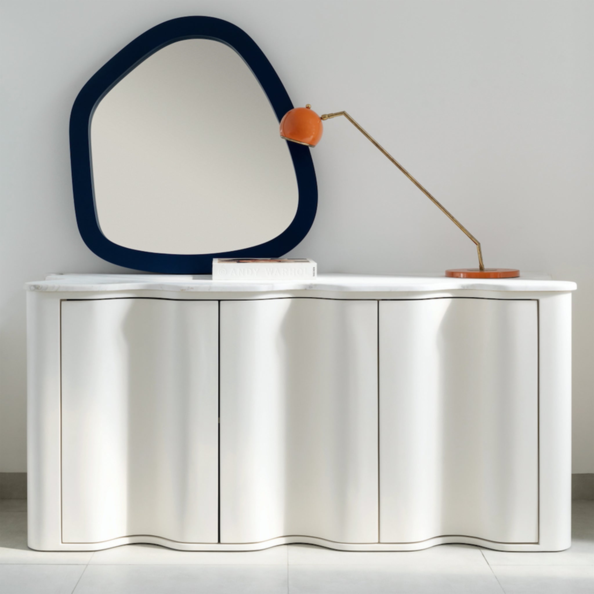 A modern sideboard with a wavy front design and a white marble top, decorated with an abstract mirror, an orange desk lamp, and a book. The sideboard is placed against a light gray wall, showcasing a stylish and contemporary aesthetic.