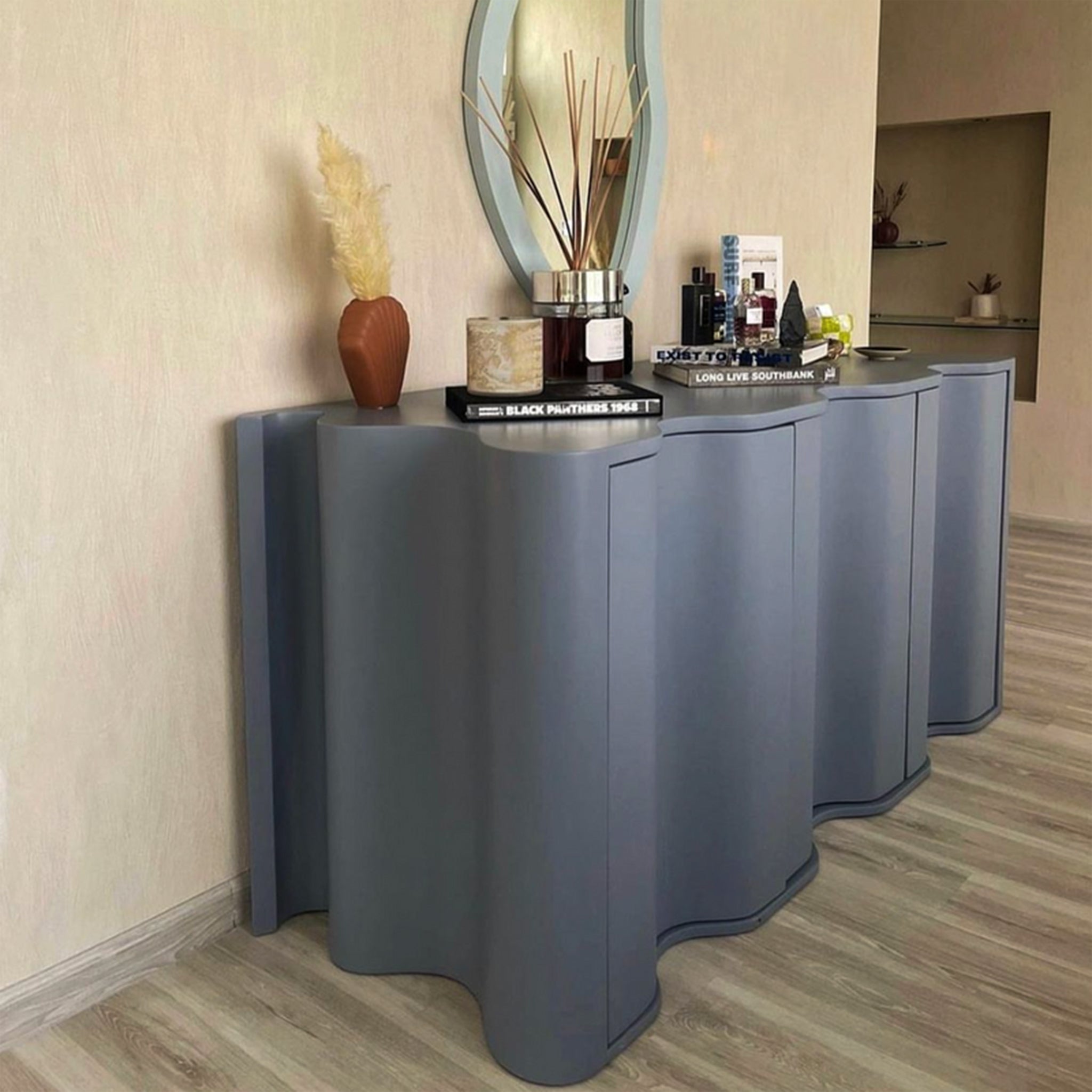 A contemporary sideboard with a wavy front design in a muted gray color, topped with various decorative items including books, a reed diffuser, and a vase with dried pampas grass. The sideboard is set against a beige wall with light wooden flooring, creating a modern and stylish interior.