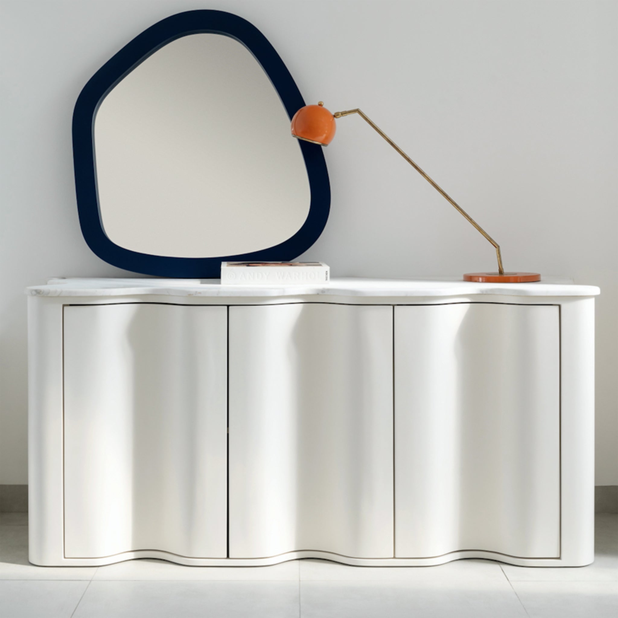 A modern sideboard with a wavy front design and a white marble top, decorated with an abstract mirror featuring a blue frame, an orange desk lamp, and a book. The sideboard is placed against a light gray wall, exemplifying a chic and contemporary interior style.