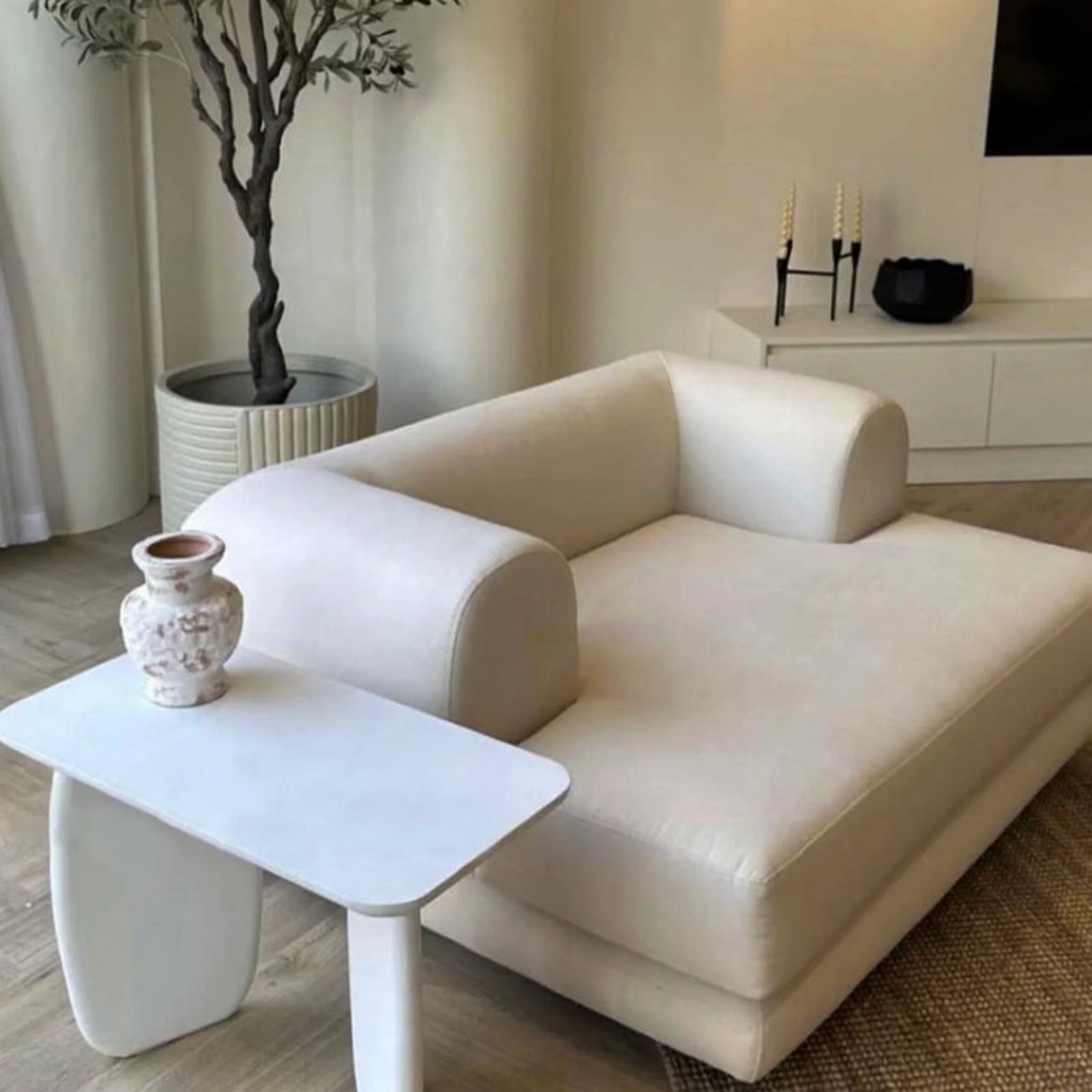 "Modern living room featuring The Lewis Low Lounger Accent Chair with beige upholstery, accompanied by a white side table holding a decorative vase, and complemented by a potted plant and minimalist decor."