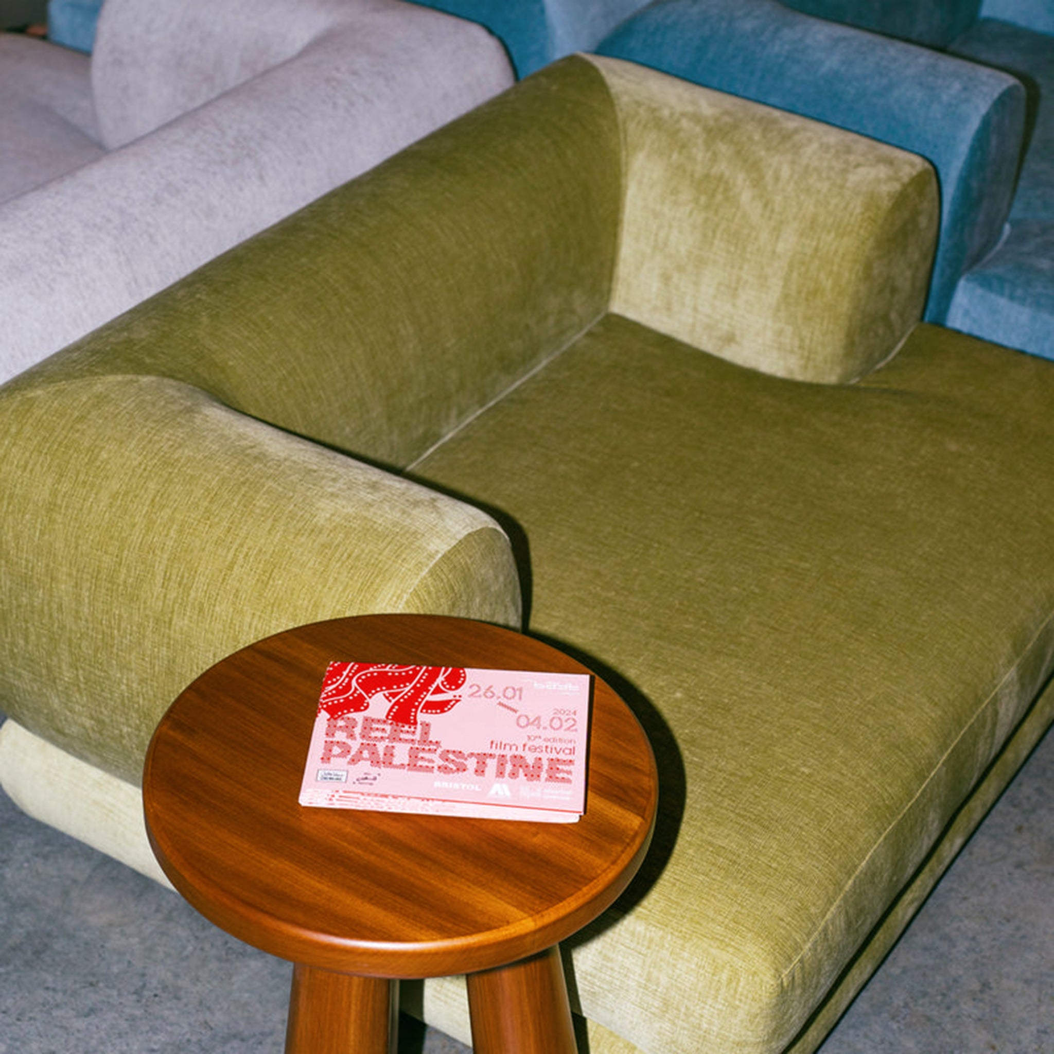 "Comfortable seating area featuring The Lewis Low Lounger Accent Chair with green upholstery, positioned next to a round wooden side table holding a pamphlet, surrounded by other colorful lounge chairs."