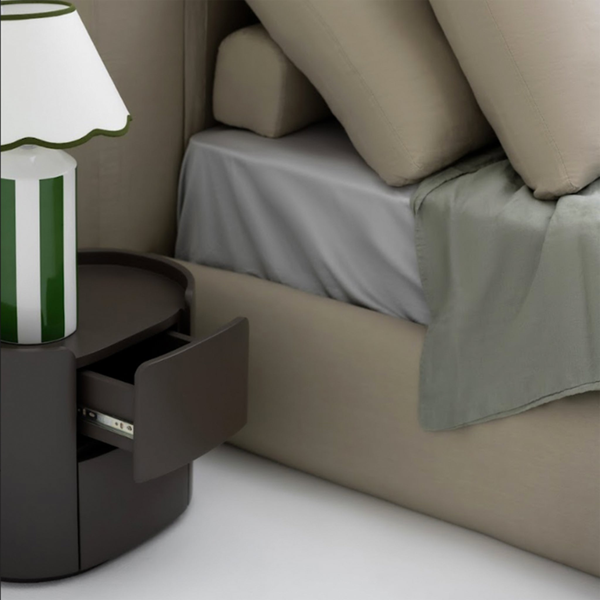 Close-up of a black cylindrical bedside table with an open drawer, next to a bed with beige bedding and gray sheets. A green and white striped table lamp sits on top of the table.