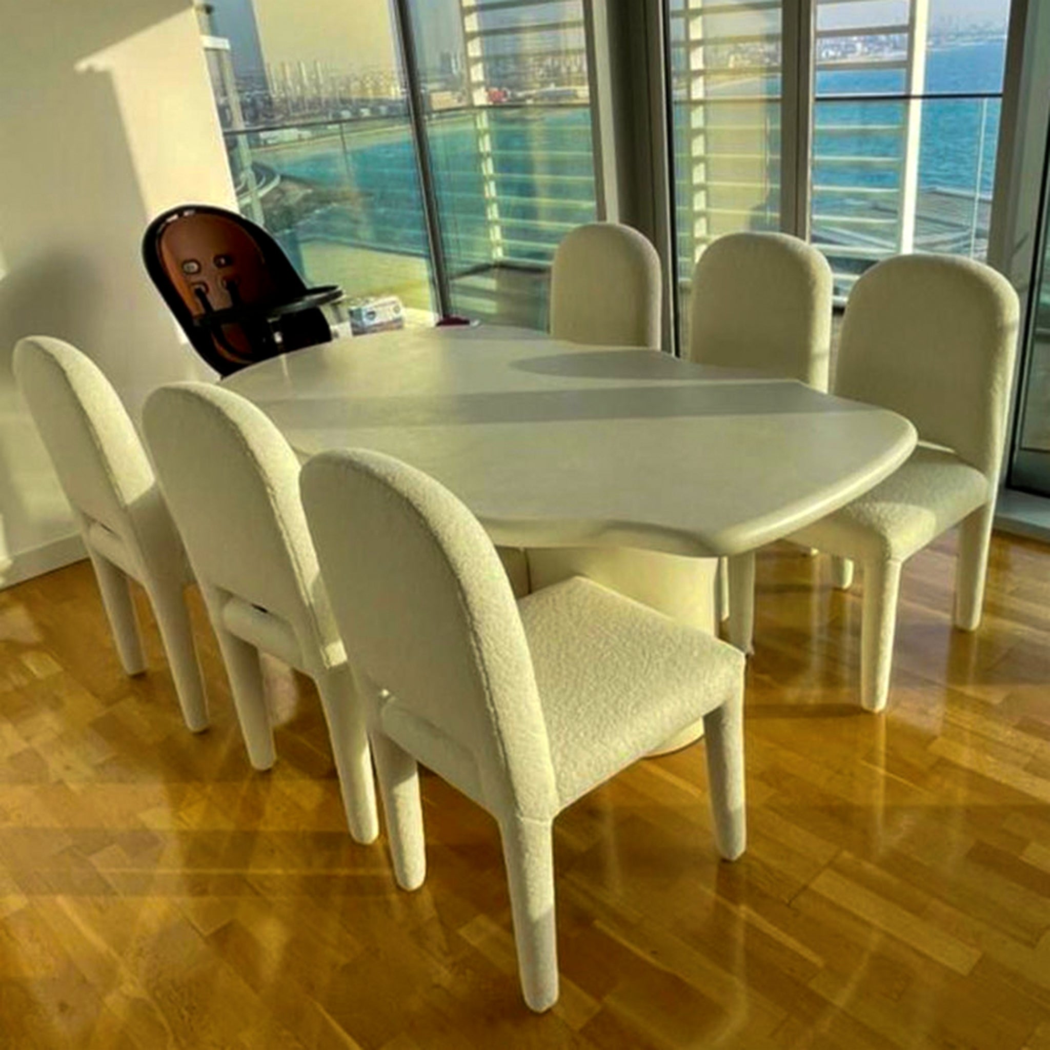 The Shelly Chair set - a modern dining table surrounded by six upholstered dining chairs with curved backs and elegant thin legs, set in a room with a view of the sea.