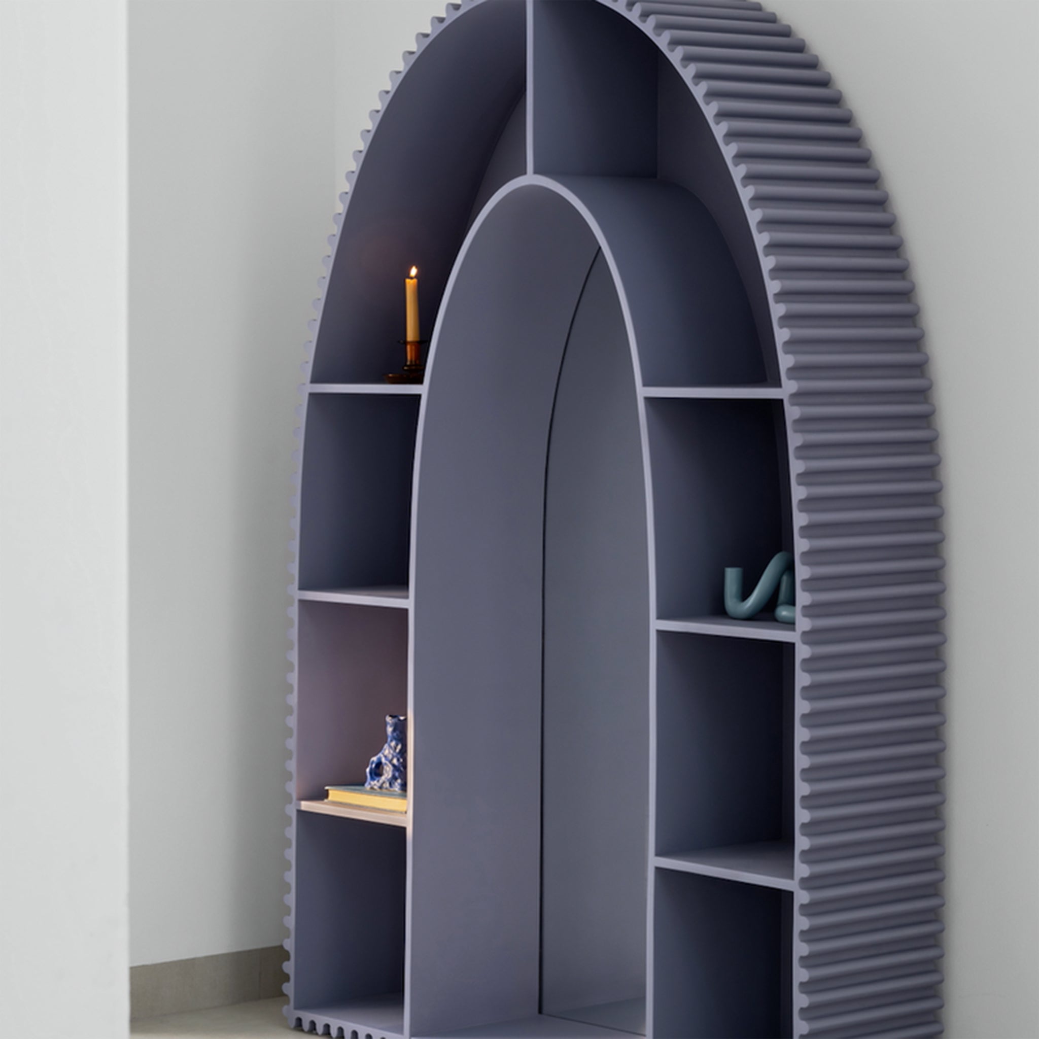 Side view of a modern, arch-shaped bookshelf in muted gray, showcasing its grooved edges and integrated mirror in the center. The shelves contain a candle, a decorative figurine, and other small items, positioned against a light gray wall.