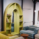 Load image into Gallery viewer, A contemporary, arch-shaped bookshelf in a vibrant yellow color with integrated mirror, situated in a modern living space. The bookshelf features grooved edges and contains books and decorative items. The area includes comfortable blue and beige sofas and a small wooden table, creating a cozy and stylish ambiance. Buy Bookshelf Dubai
