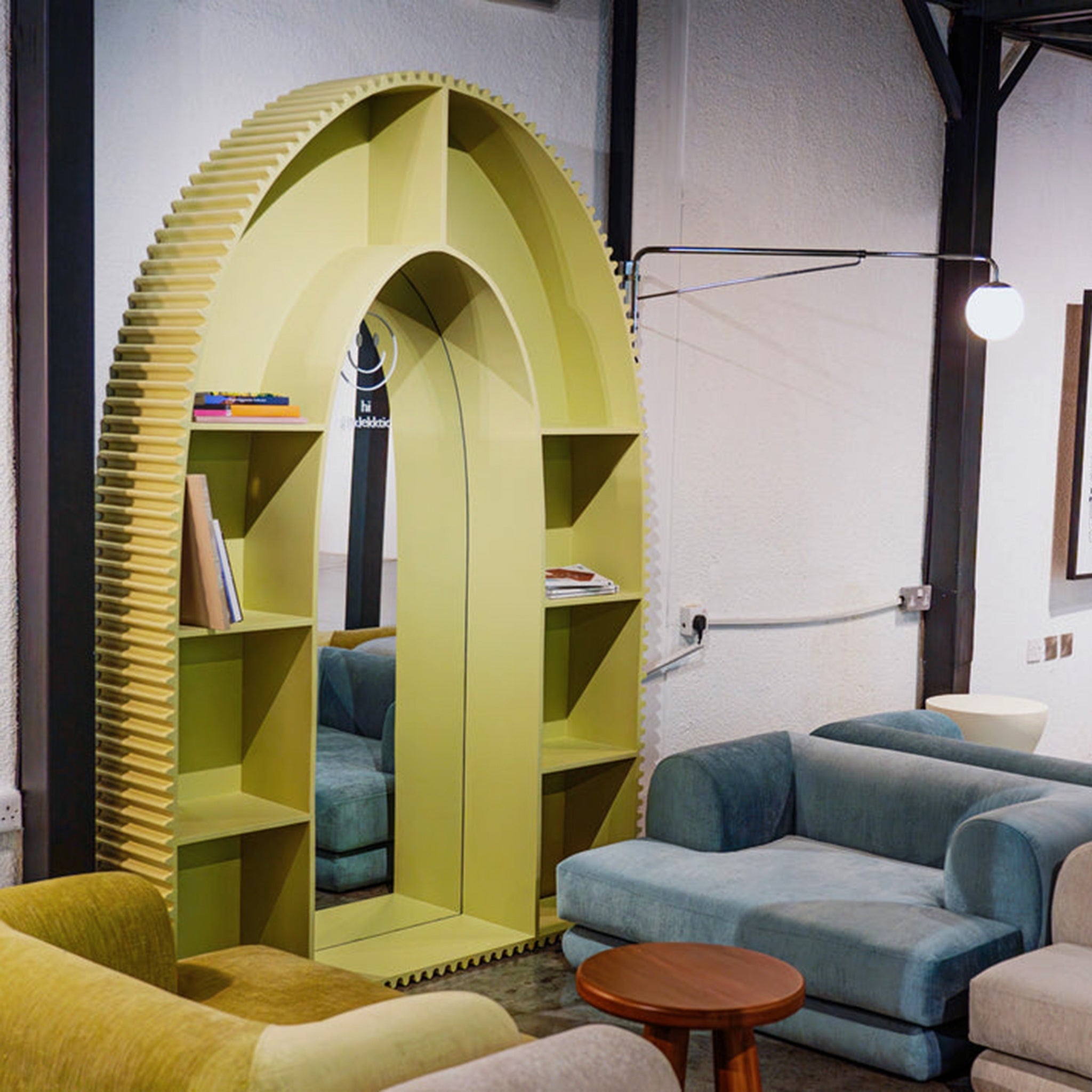 A contemporary, arch-shaped bookshelf in a vibrant yellow color with integrated mirror, situated in a modern living space. The bookshelf features grooved edges and contains books and decorative items. The area includes comfortable blue and beige sofas and a small wooden table, creating a cozy and stylish ambiance.