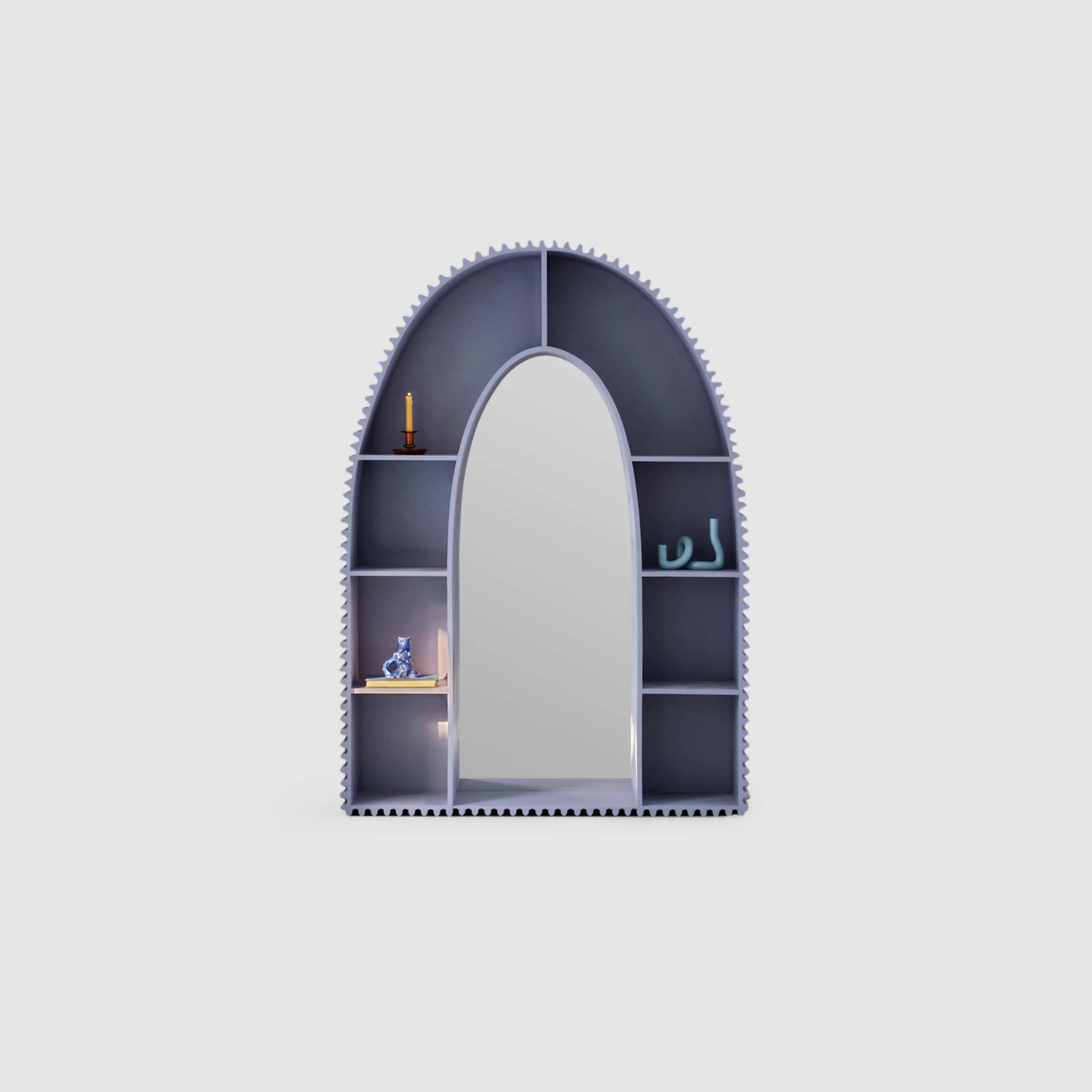 A modern, arch-shaped bookshelf in muted gray with integrated mirror in the center, surrounded by small compartments containing a candle, a decorative figurine, and other small items against a plain white background.