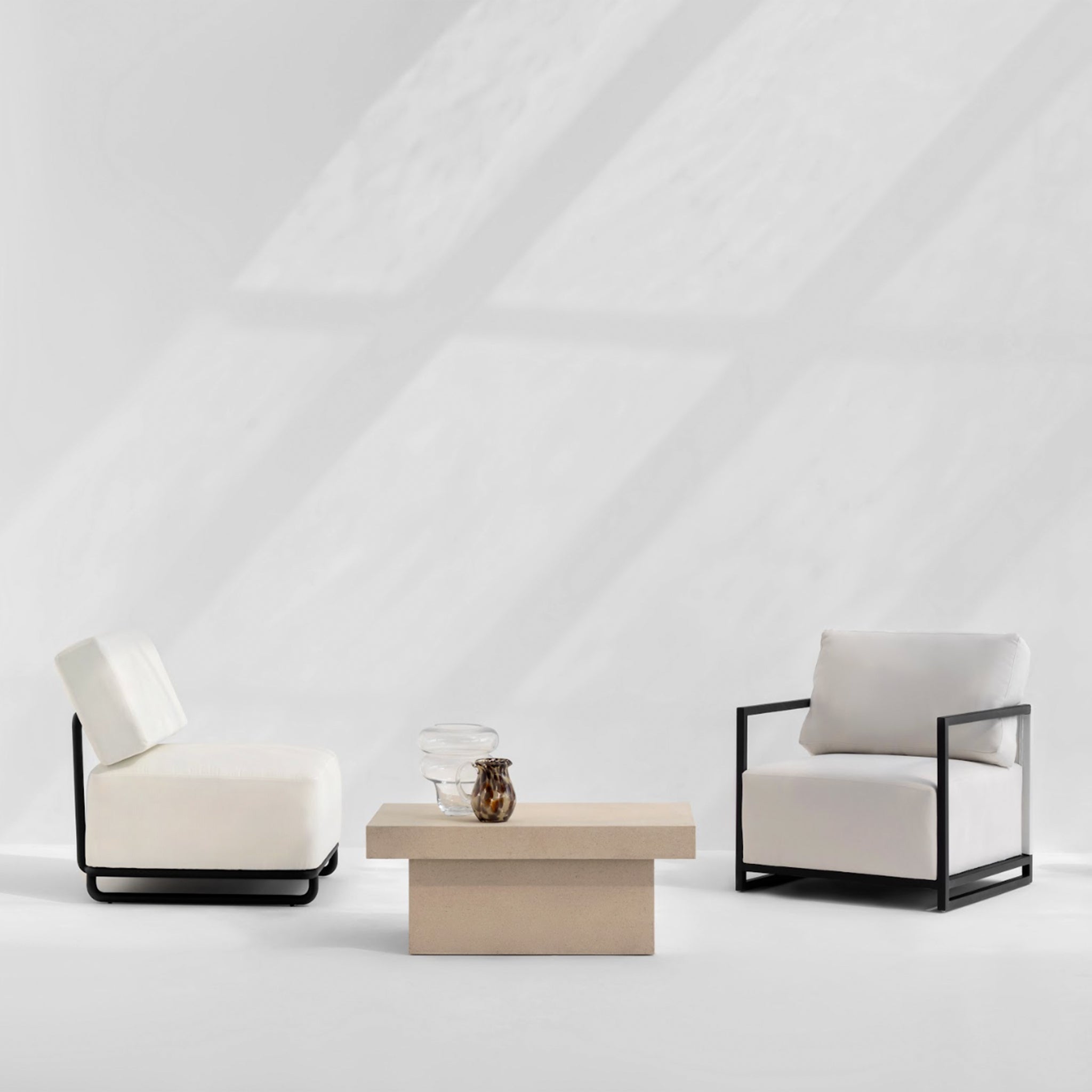Modern outdoor seating arrangement featuring two Wyth Outdoor Accent Chairs with white cushions and black metal frames, accompanied by a minimalist beige coffee table with decorative glassware.