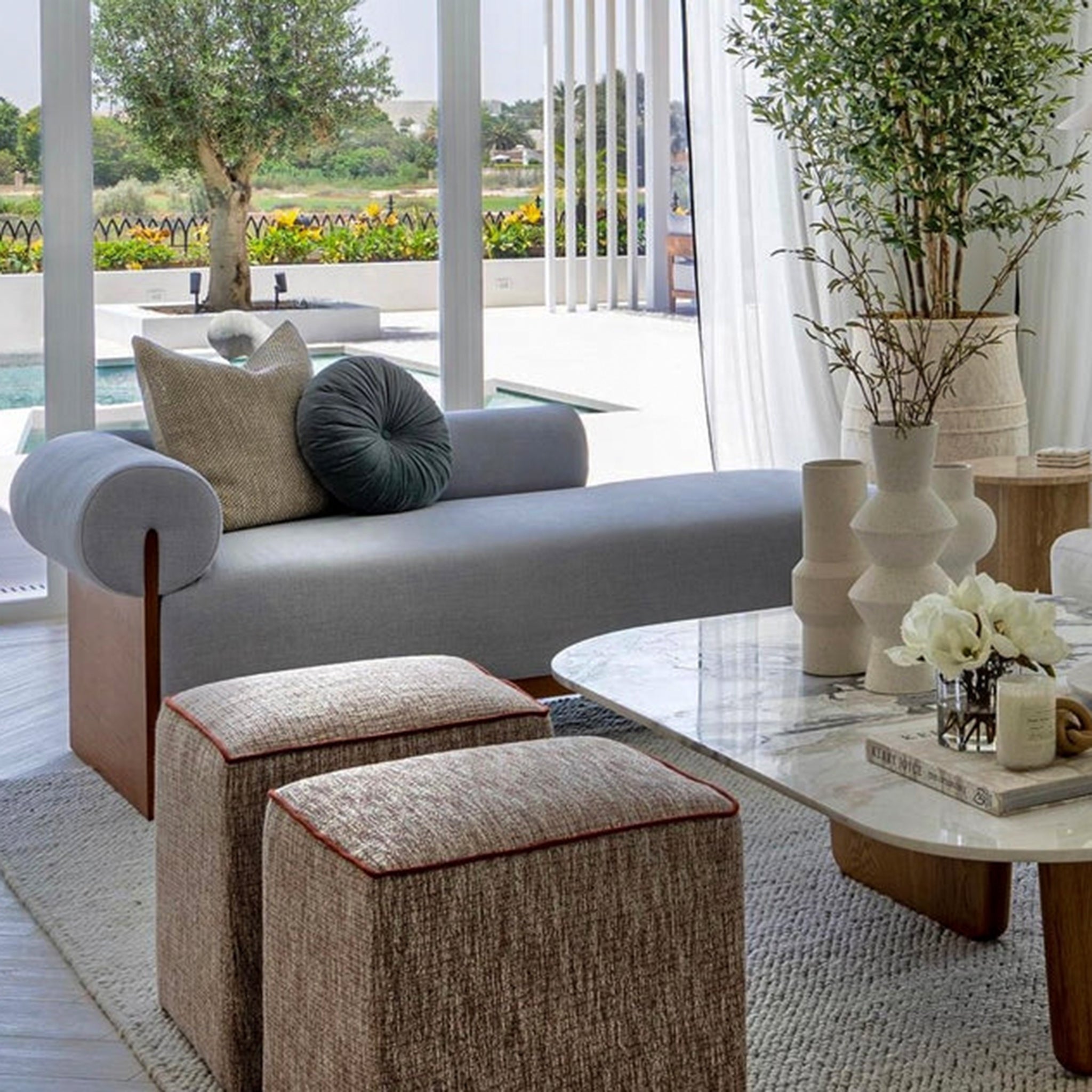 The Devon Chaise in a modern living room with a grey upholstered seat and wooden frame, positioned by large windows overlooking a garden and pool, complemented by decorative pillows and a stylish coffee table with vases and flowers.